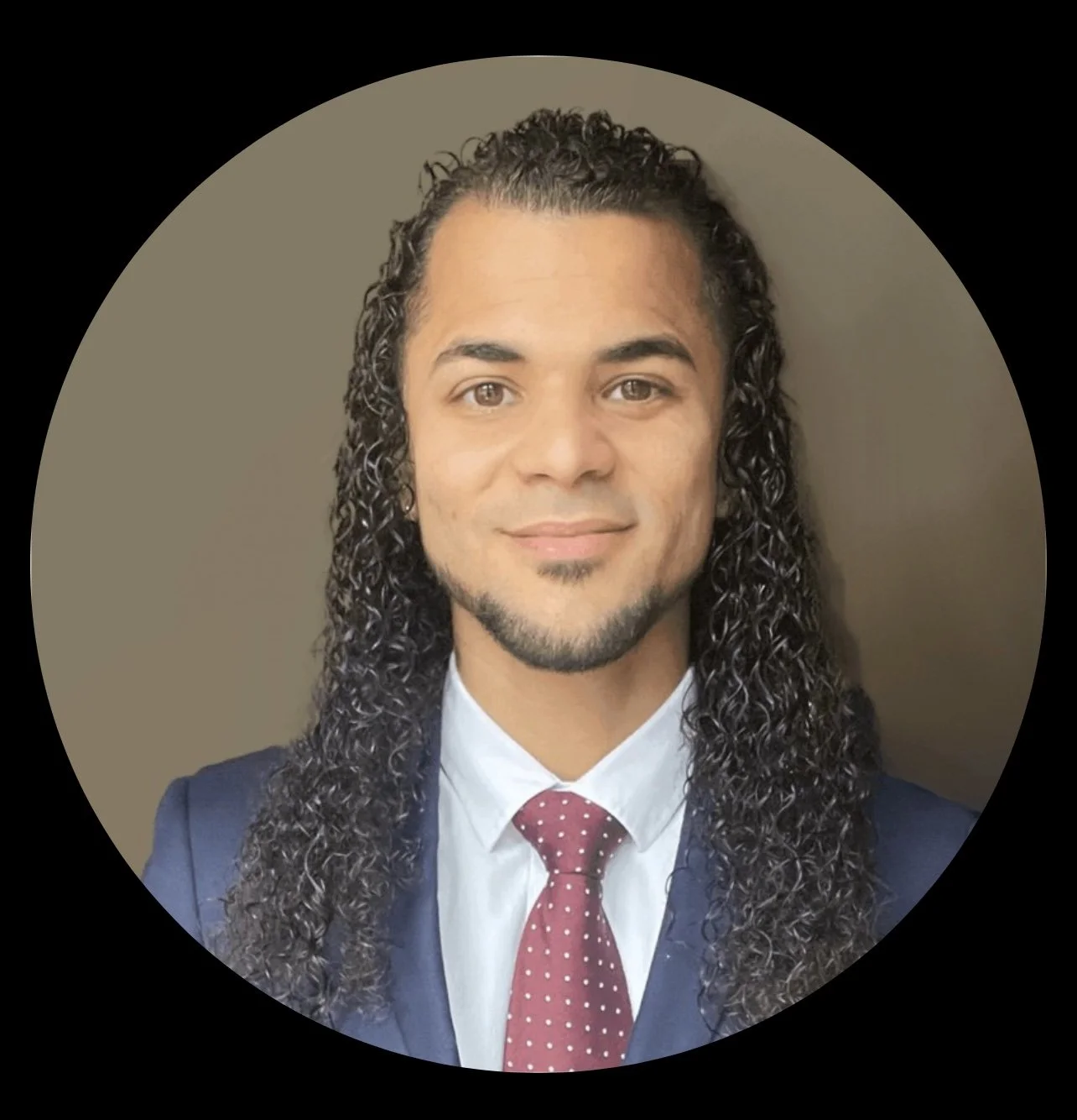 Man with long curly hair in a blue suit and red polka dot tie, smiling in a portrait.