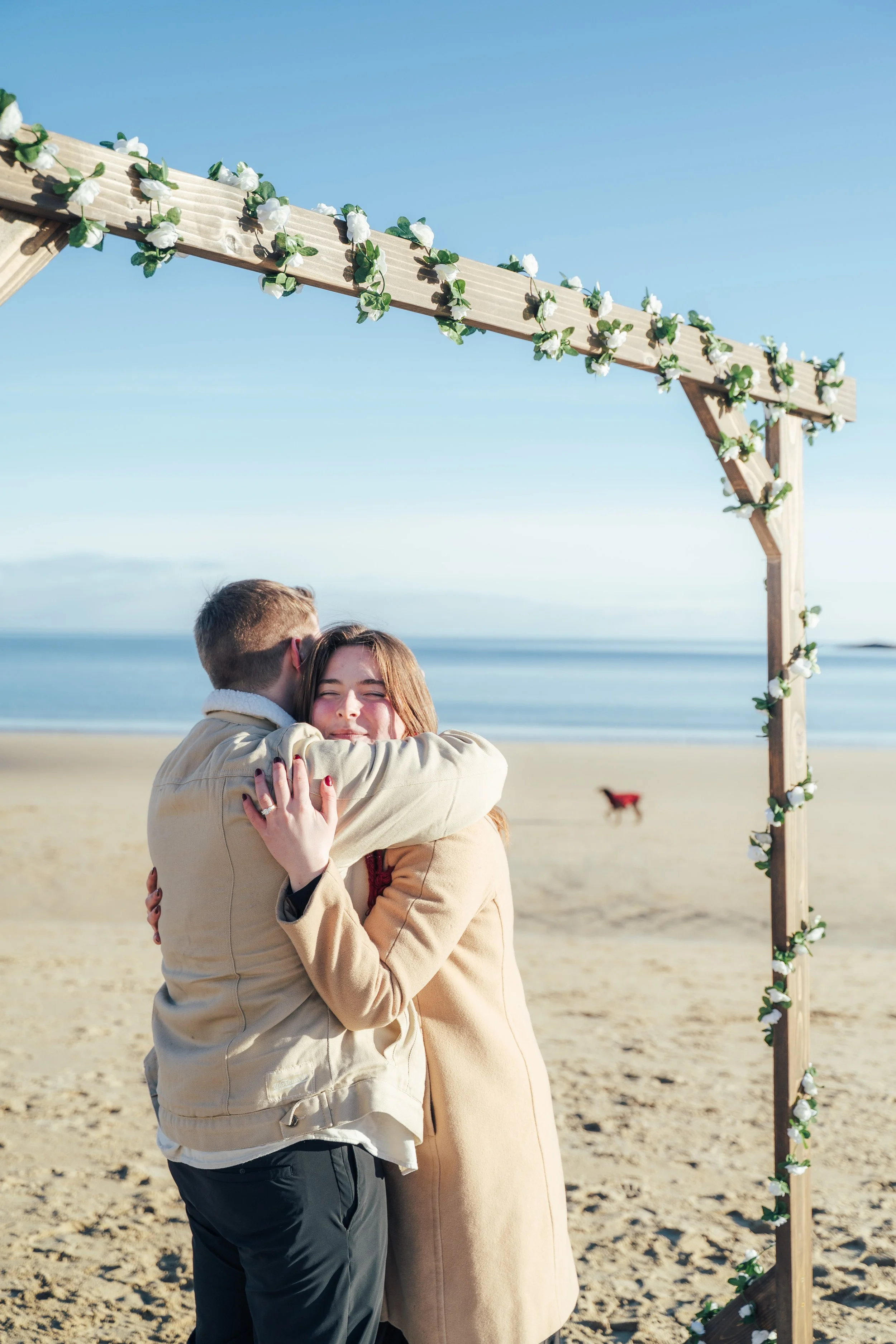 A couple hugging on a beach during an engagement with an arch decorated with flowers.