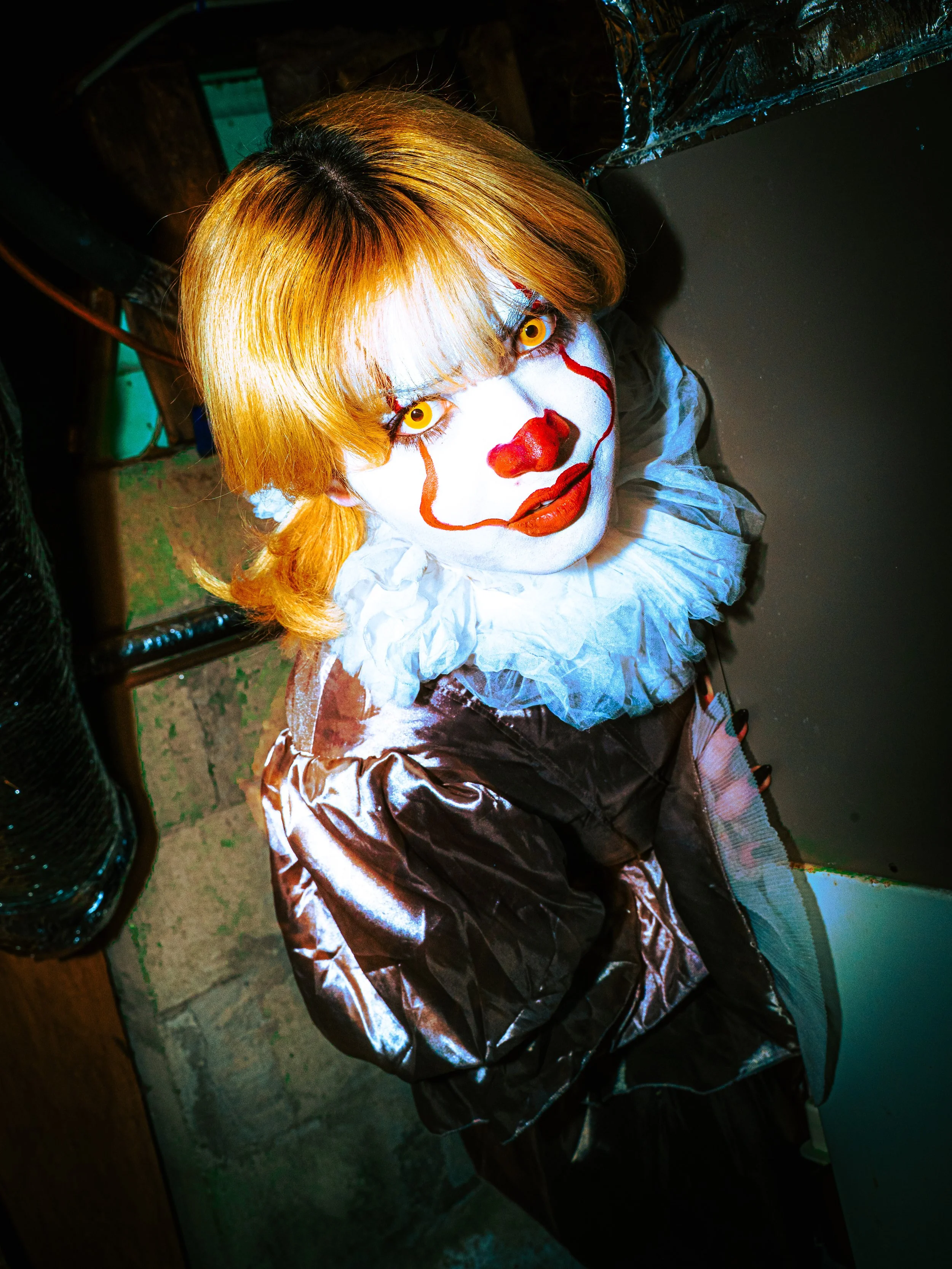 A person dressed as Pennywise the clown from It, with orange hair, white face paint, red nose, yellow eyes, and red makeup details, sitting at a table.