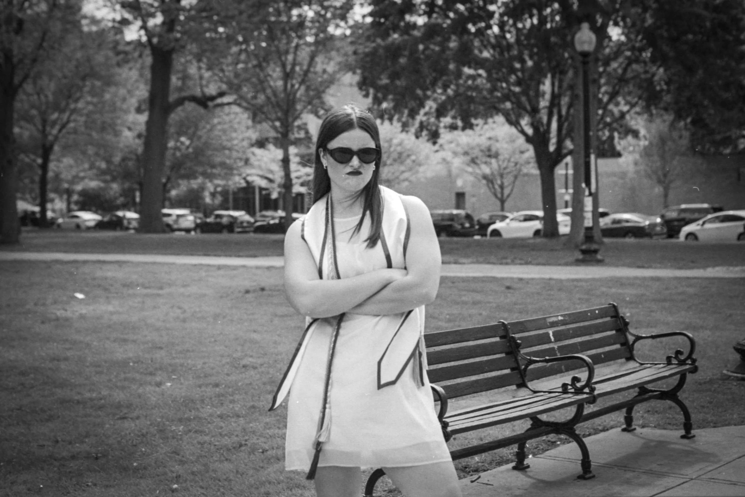 A woman with long dark hair, wearing sunglasses, a sleeveless top, and a dress, stands with crossed arms in a park with benches, trees, and parked cars in the background.