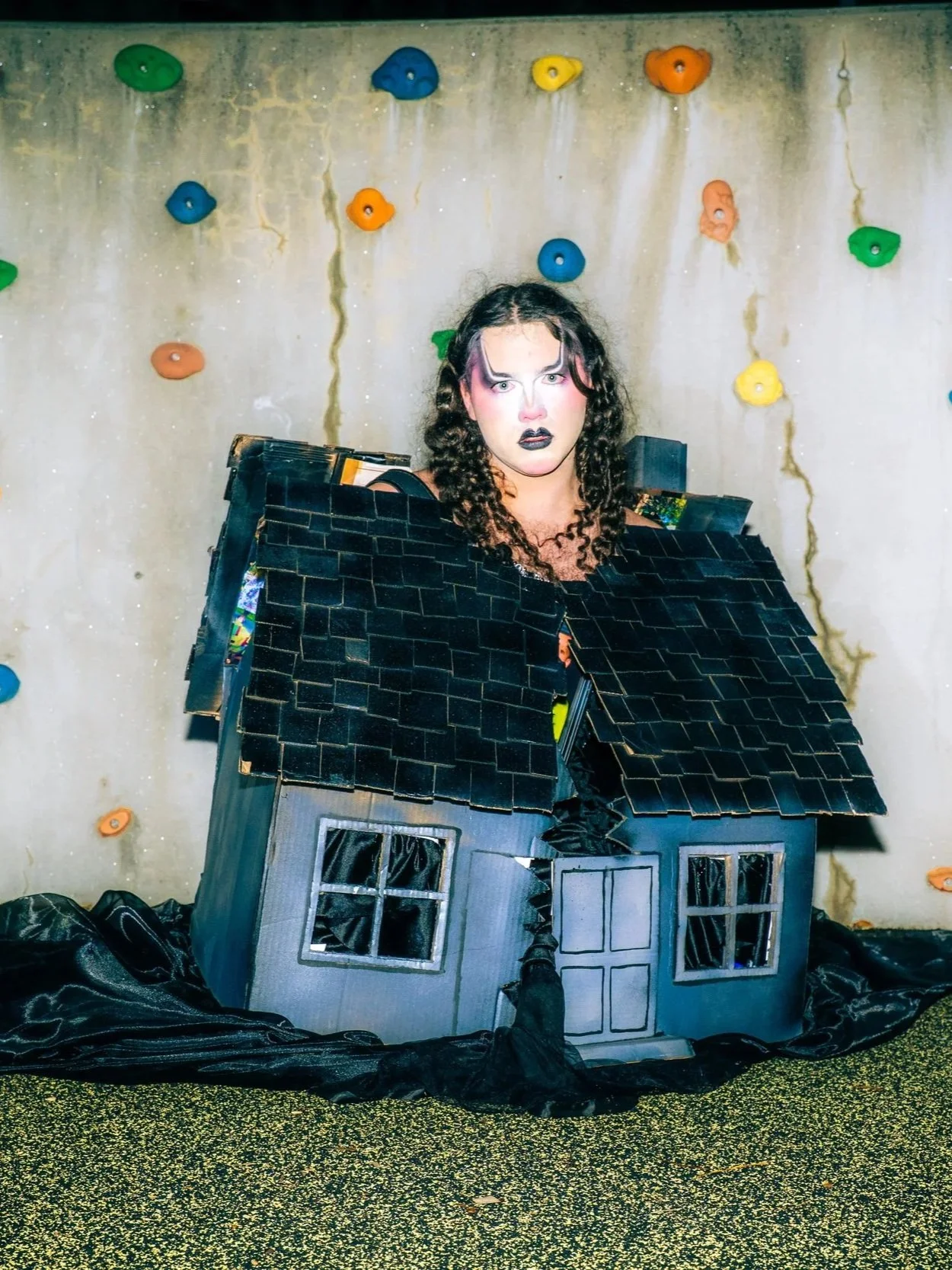 Person dressed in a black costume that resembles a damaged house, with black hair and gothic makeup, standing in front of a wall with colorful climbing holds.