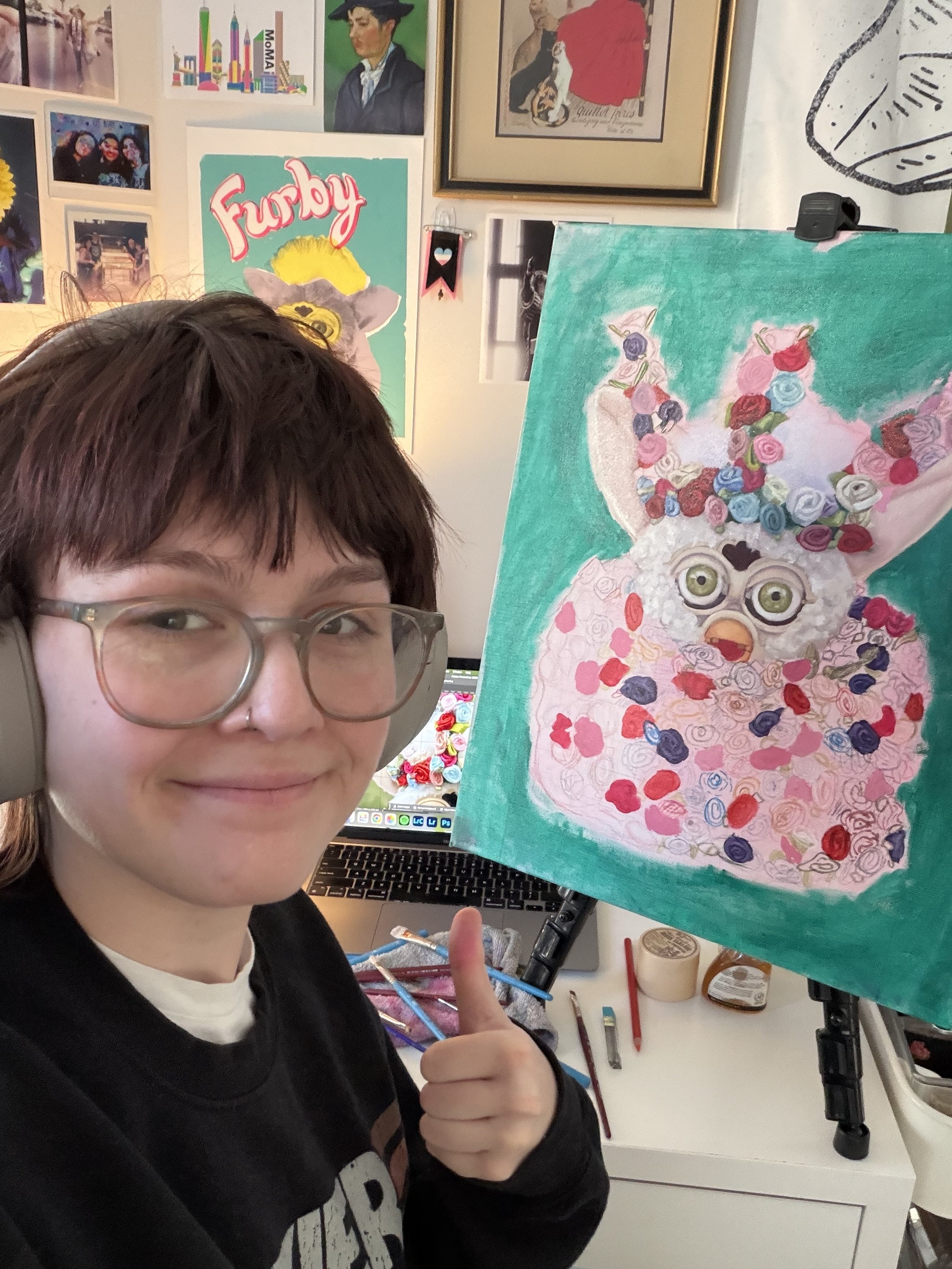 An artist with glasses and a septum piercing shows a painting of a cartoon sheep with glasses, flowers on its head, on a pink background in an art studio.