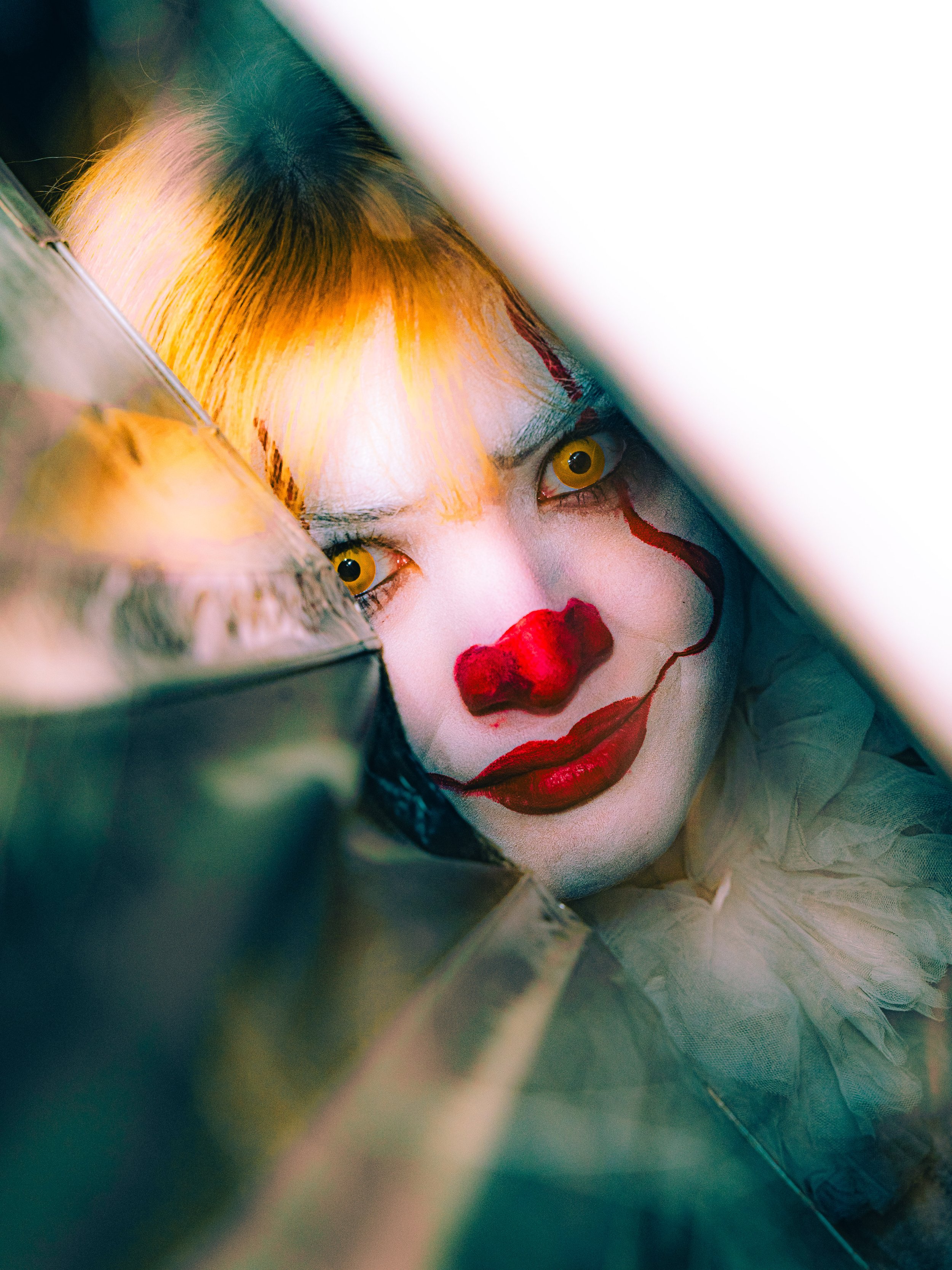 Close-up of a person dressed as a creepy clown with yellow eyes, white face paint, red clown nose, and red painted mouth, peering through a reflective surface.