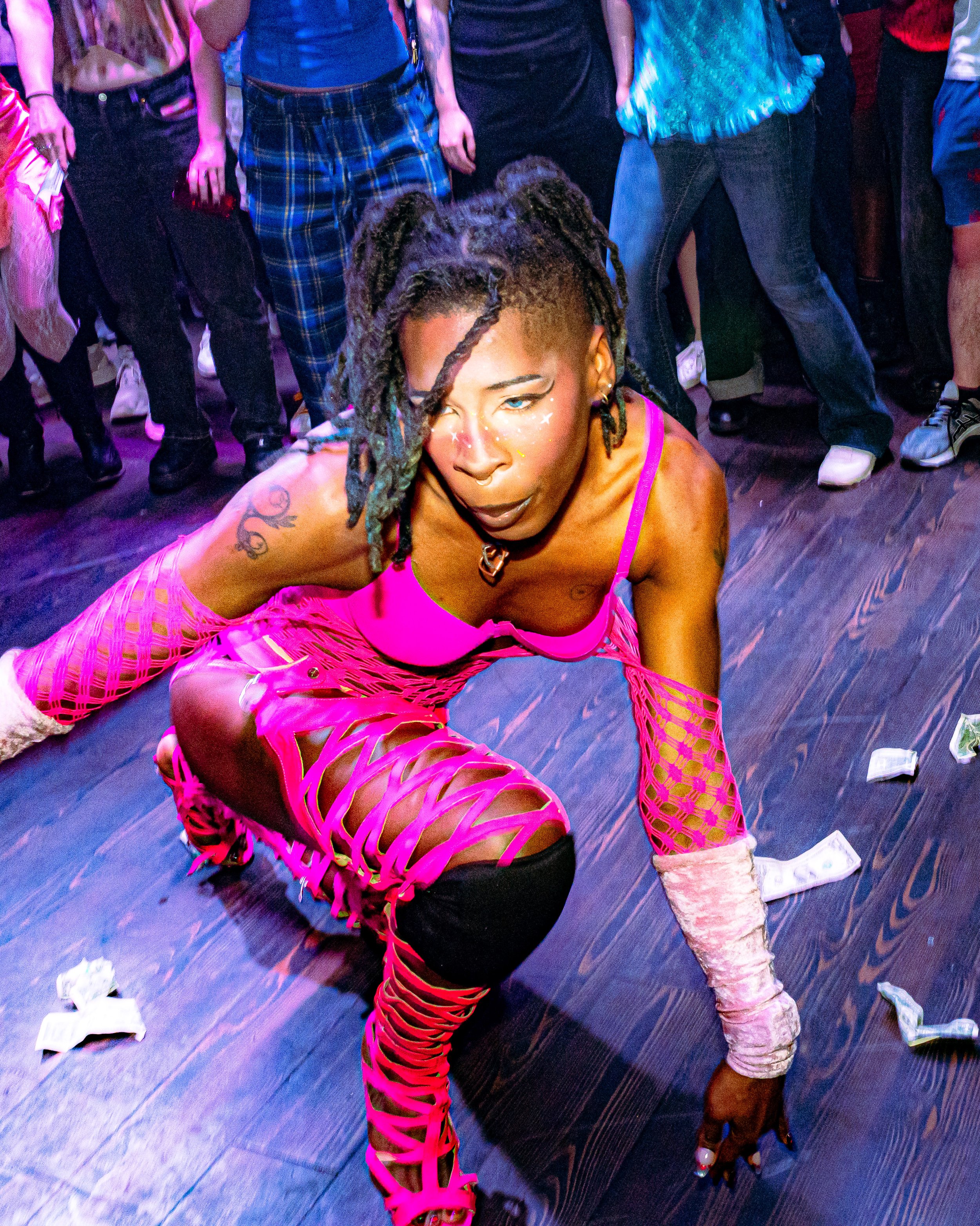 A person with dreadlocks crouches on the floor in a vibrant pink outfit with cutouts and lattice design, surrounded by a group of people at a party or dance event with dollar bills scattered on the floor.