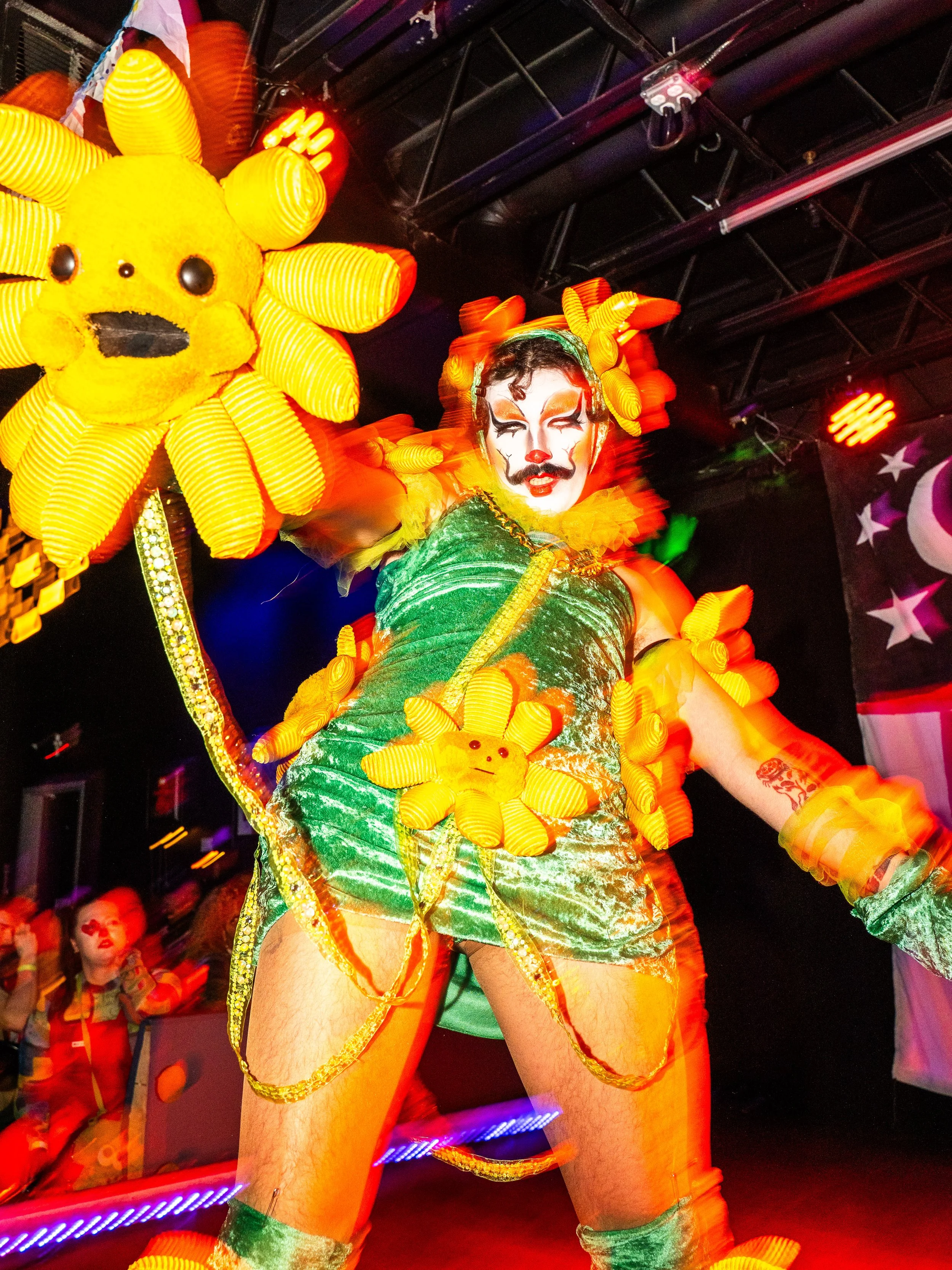 Performer dressed as a sunflower with vibrant yellow petals and a green costume, striking a pose at a neon-lit event with other performers in the background.