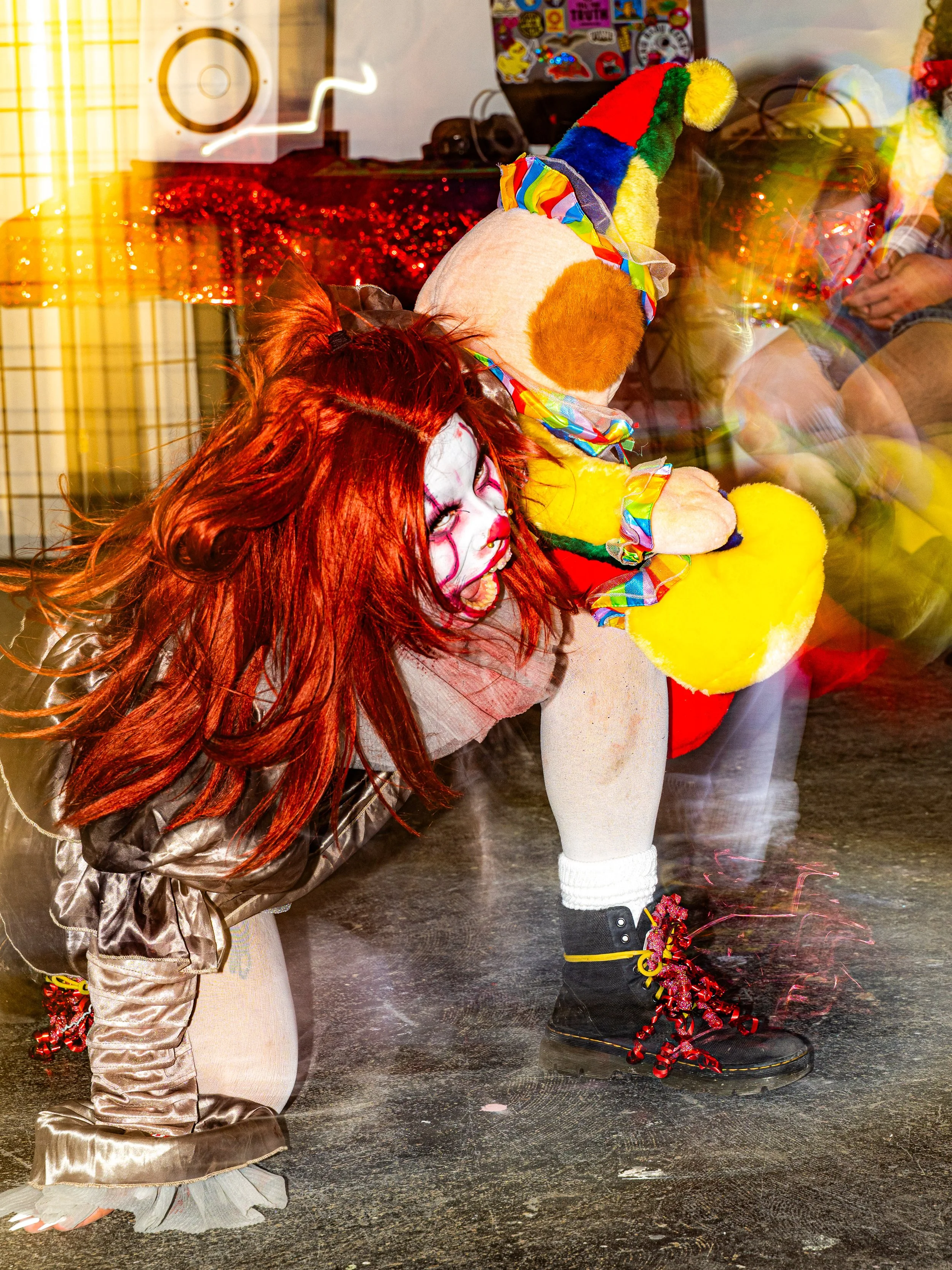 Person dressed as Pennywise the clown with red hair and white face paint, crouching on the floor, holding a plush clown doll with a rainbow hat, in a room decorated for a Halloween or costume event.