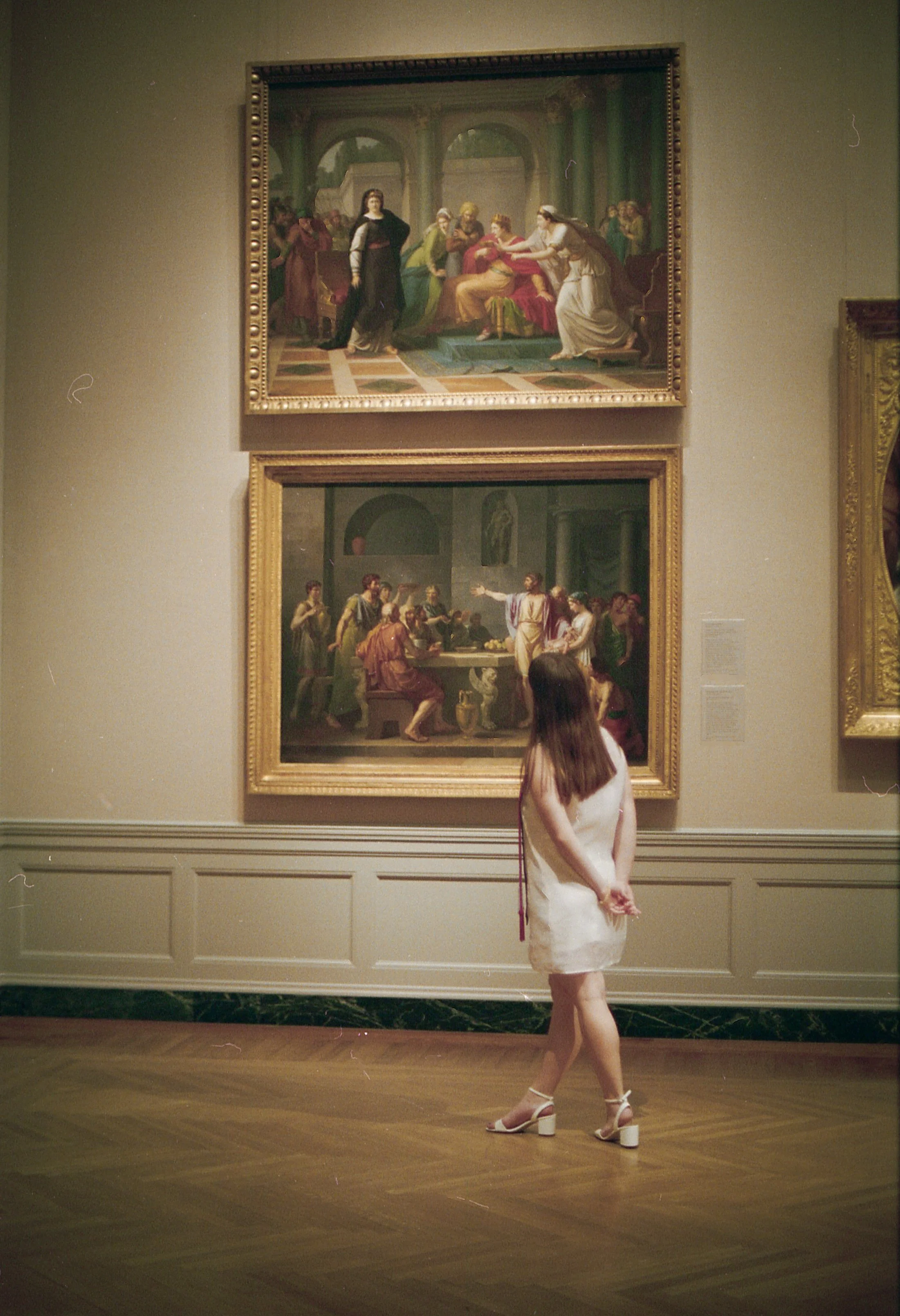 A woman in a white dress and high heels looking at two paintings in an art gallery. The paintings depict historical or mythological scenes with multiple figures.