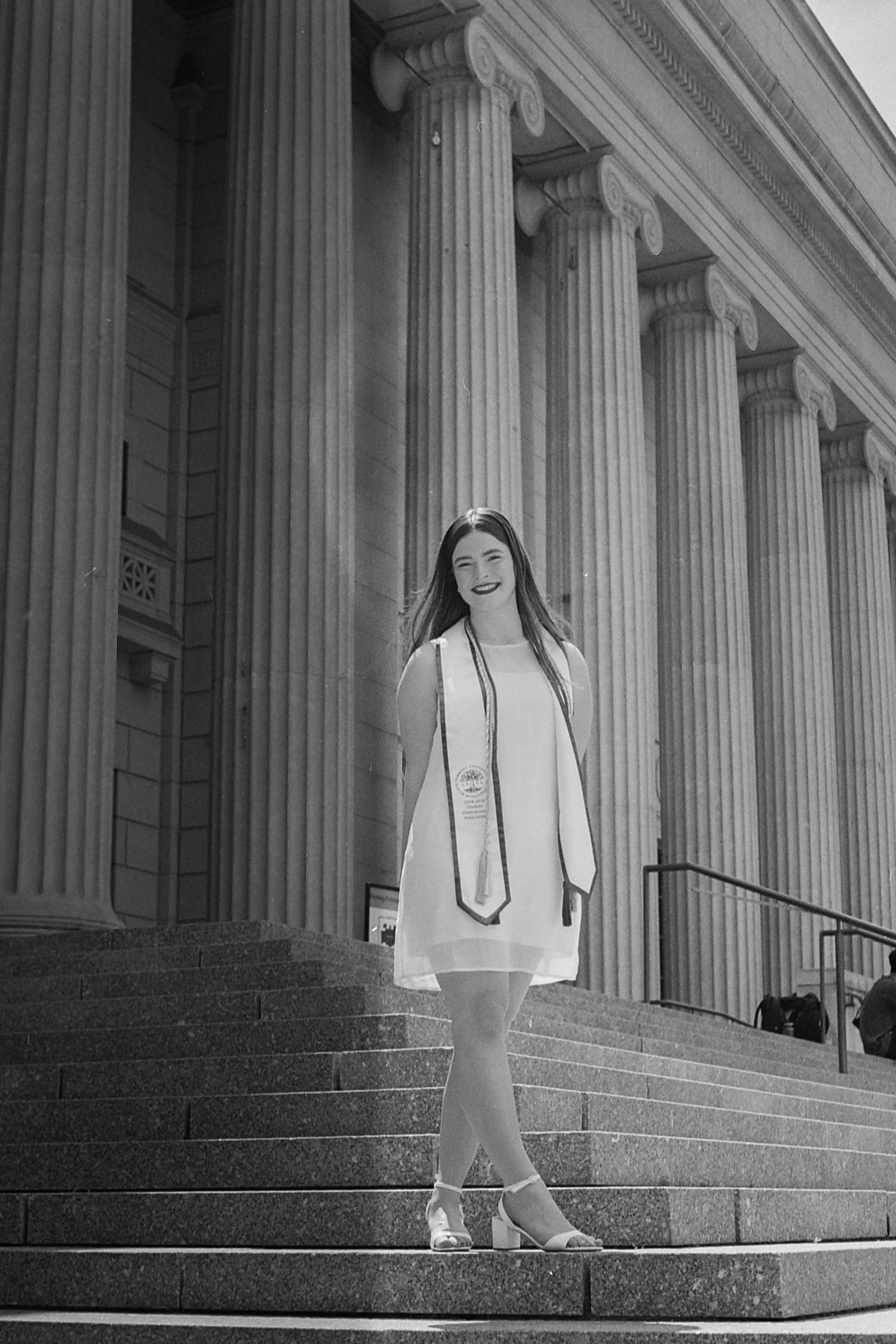 A young woman in a white dress with a graduation stole and medal, standing on steps outside a building with tall columns, smiling.