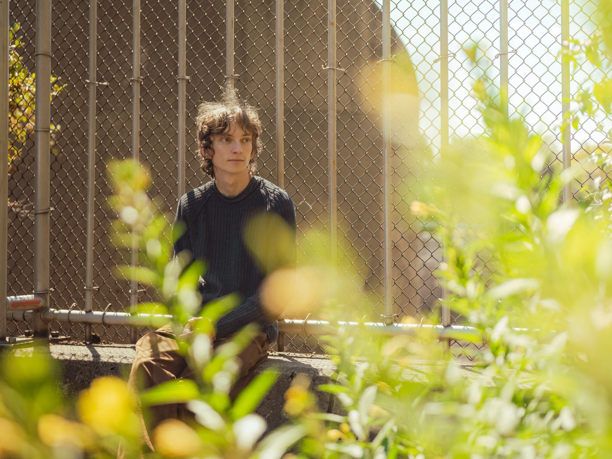 A young man with curly hair wearing a black sweater and brown pants sitting on a concrete ledge in front of a chain-link fence, surrounded by green bushes with sunlight filtering through.