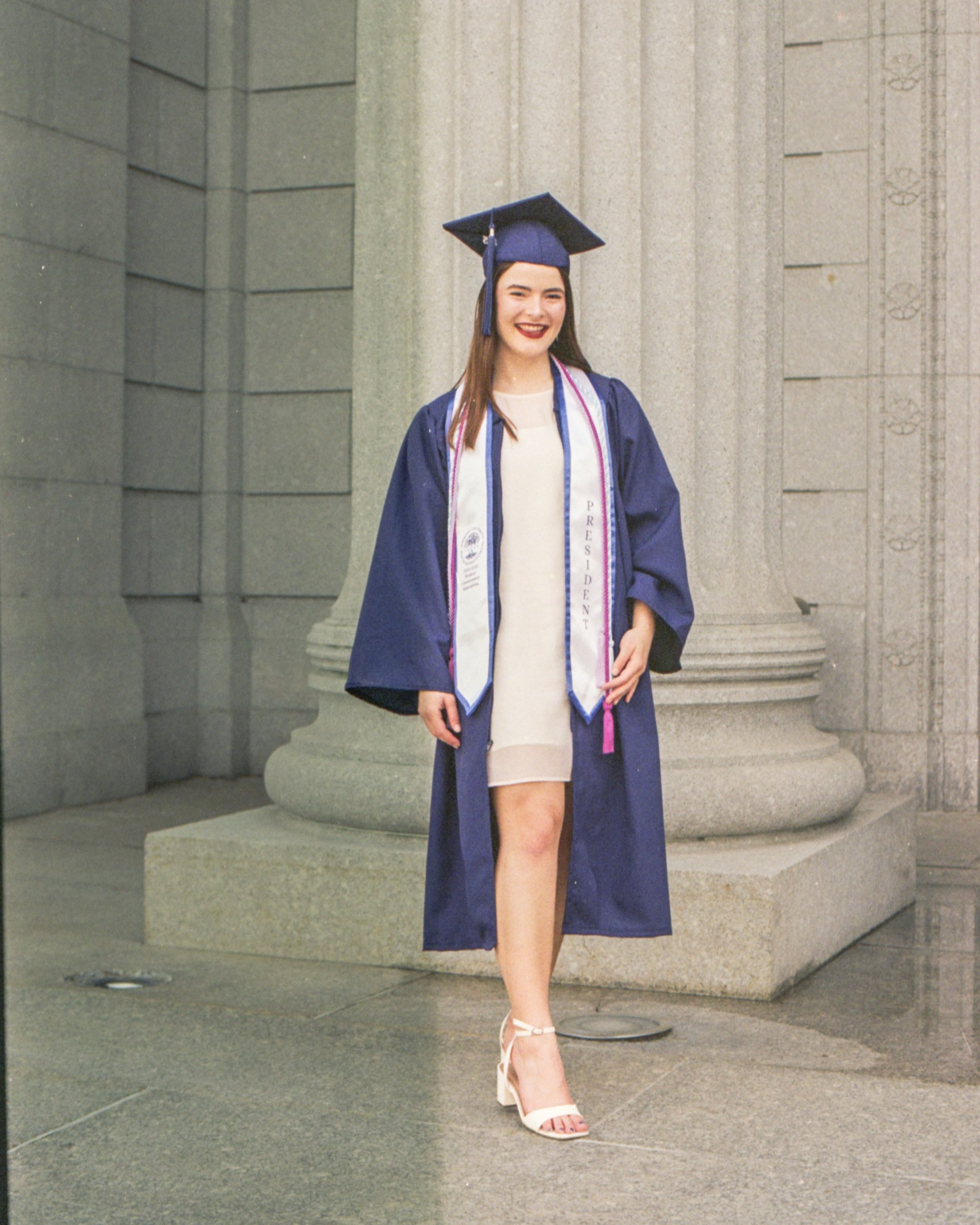 A young woman in a blue graduation gown and cap, with a white stole, standing in front of a stone building, smiling, wearing high heels.