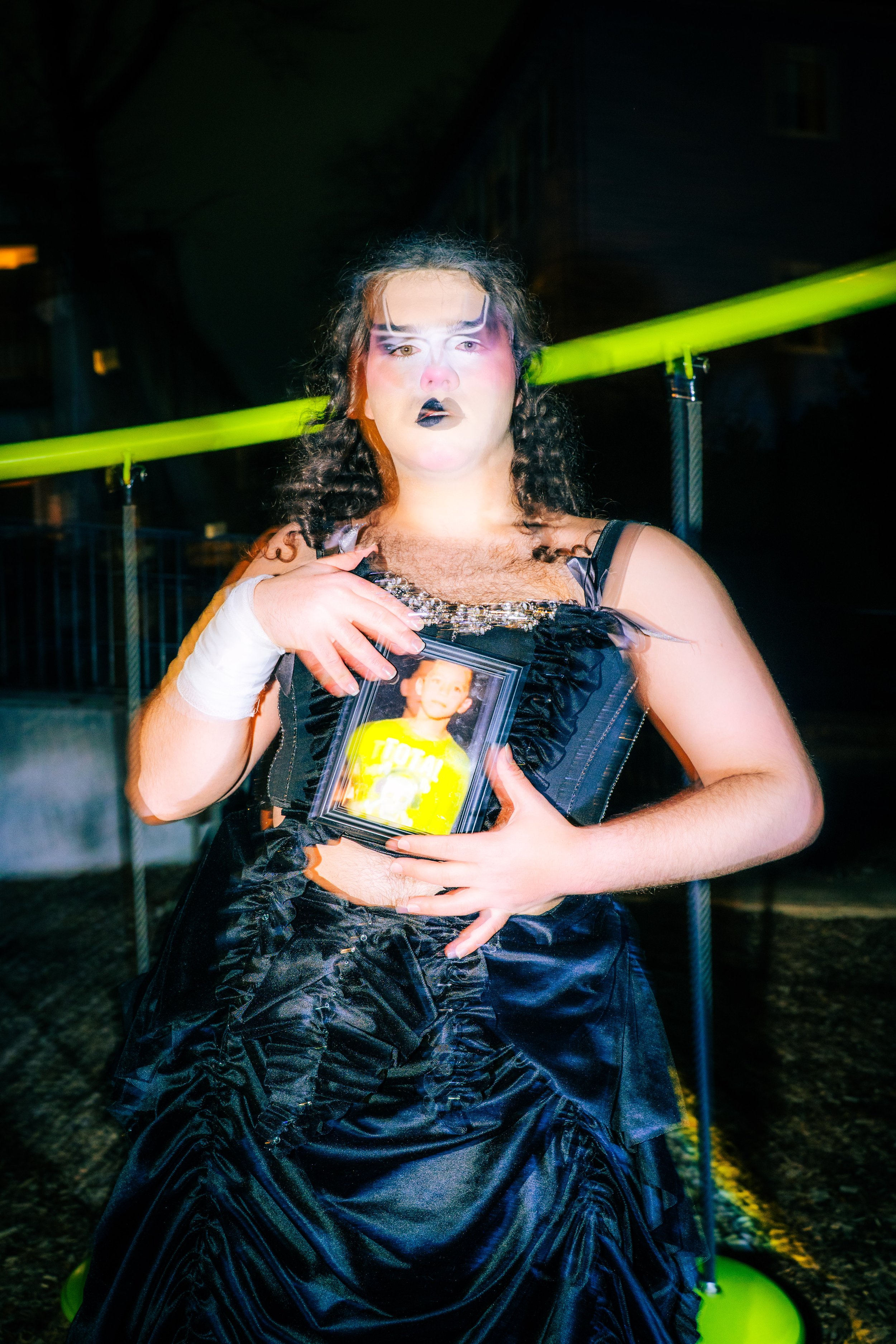Person dressed in dark, ruffled clothing with dramatic makeup holding a framed family photo in front of a green barrier at night.