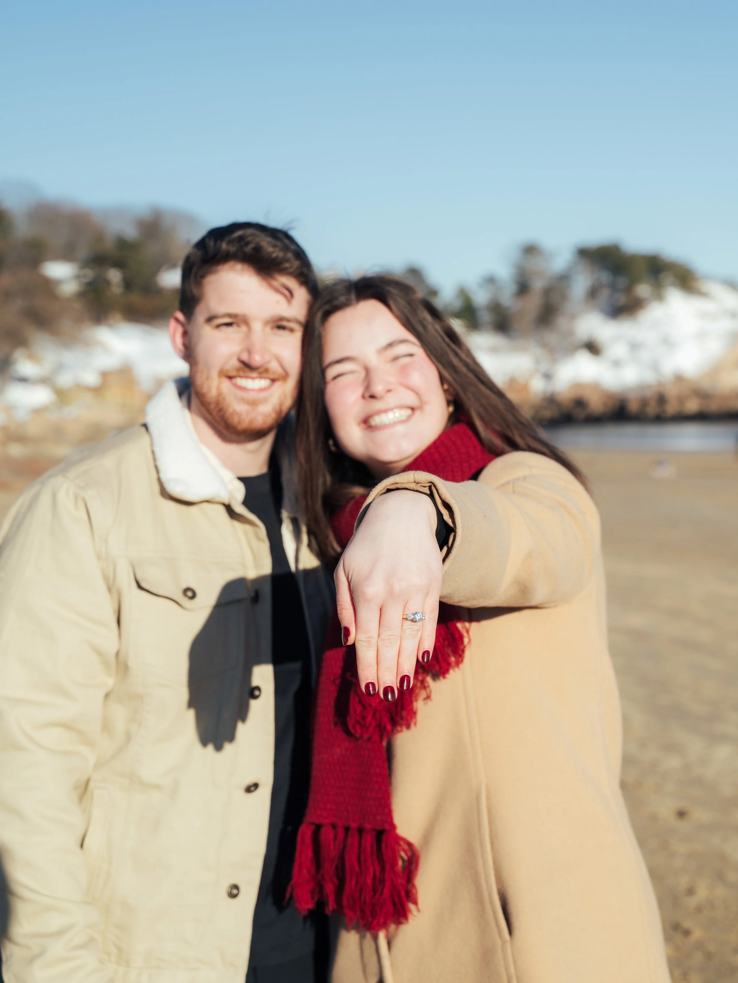 A smiling couple outdoors near the beach, with the woman displaying her engagement ring.