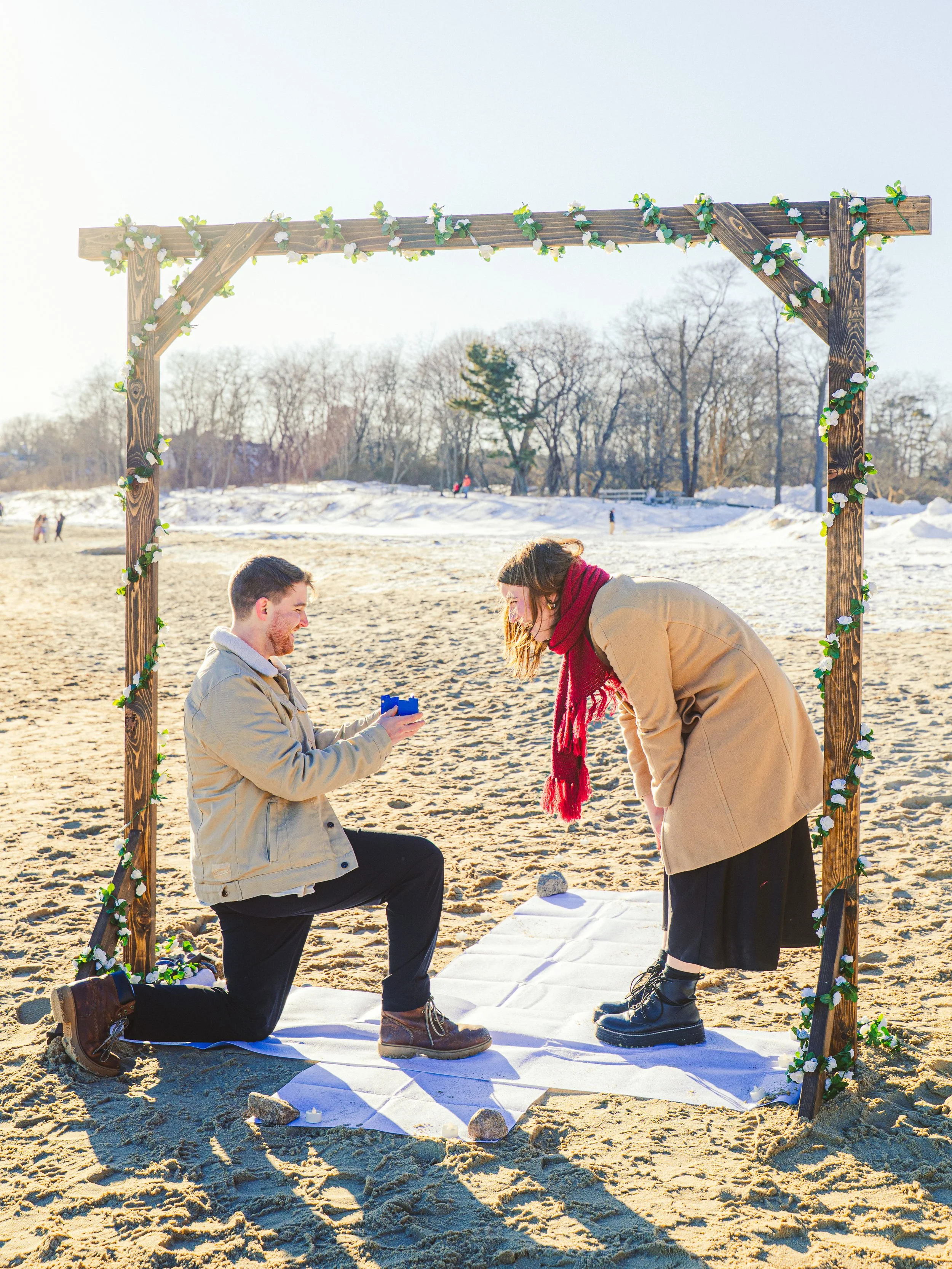 A couple is proposing on a beach in winter, under a wooden arch decorated with greenery and white flowers. The man is kneeling and holding a small box, proposing to the woman who is bending forward with joy.