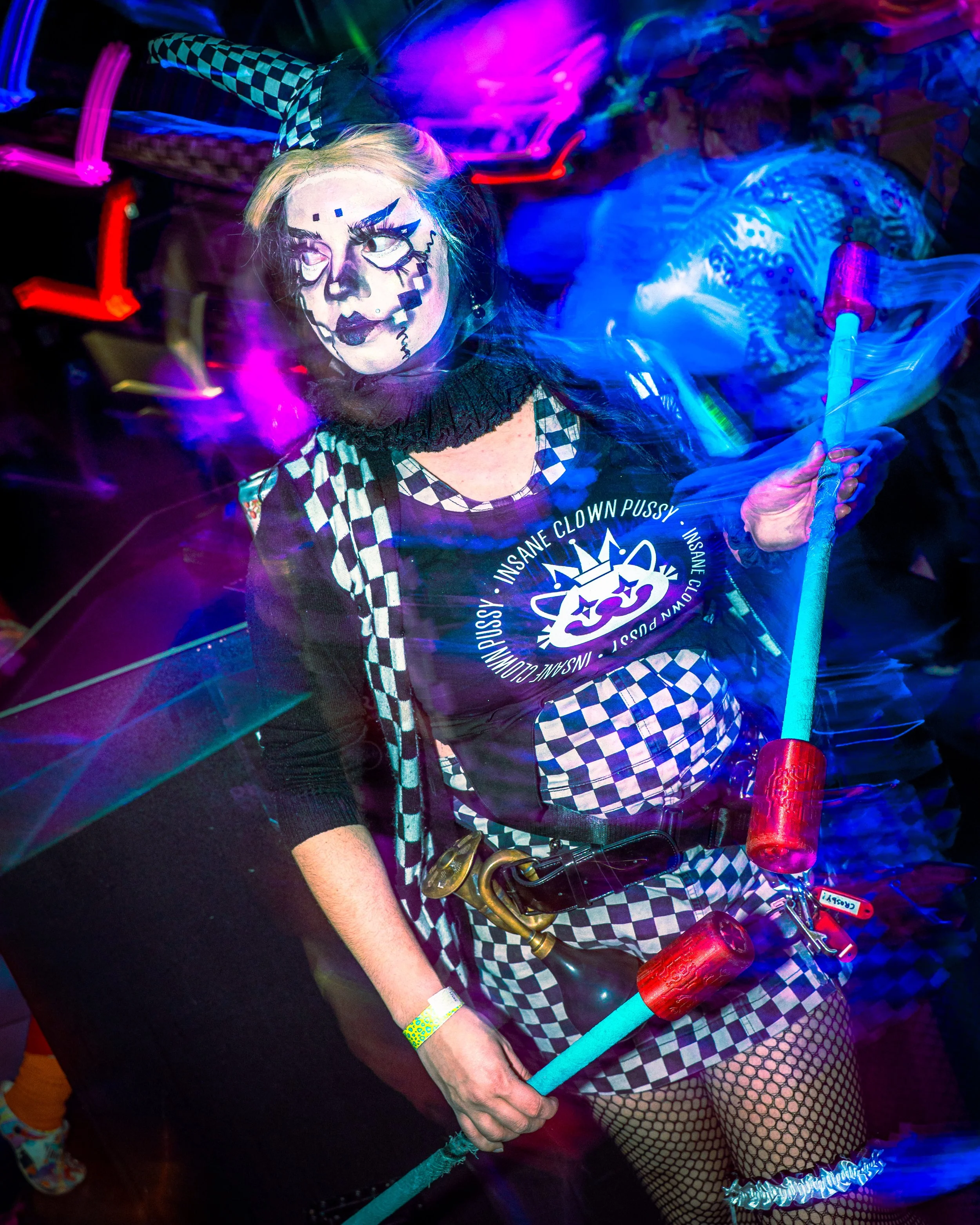 Person in punk costume with face painted as a creepy clown, wearing a 'Insane Clown Pussy' shirt, fishnet tights, and checkered clothing holding neon drumsticks at a party or event with colorful lights.