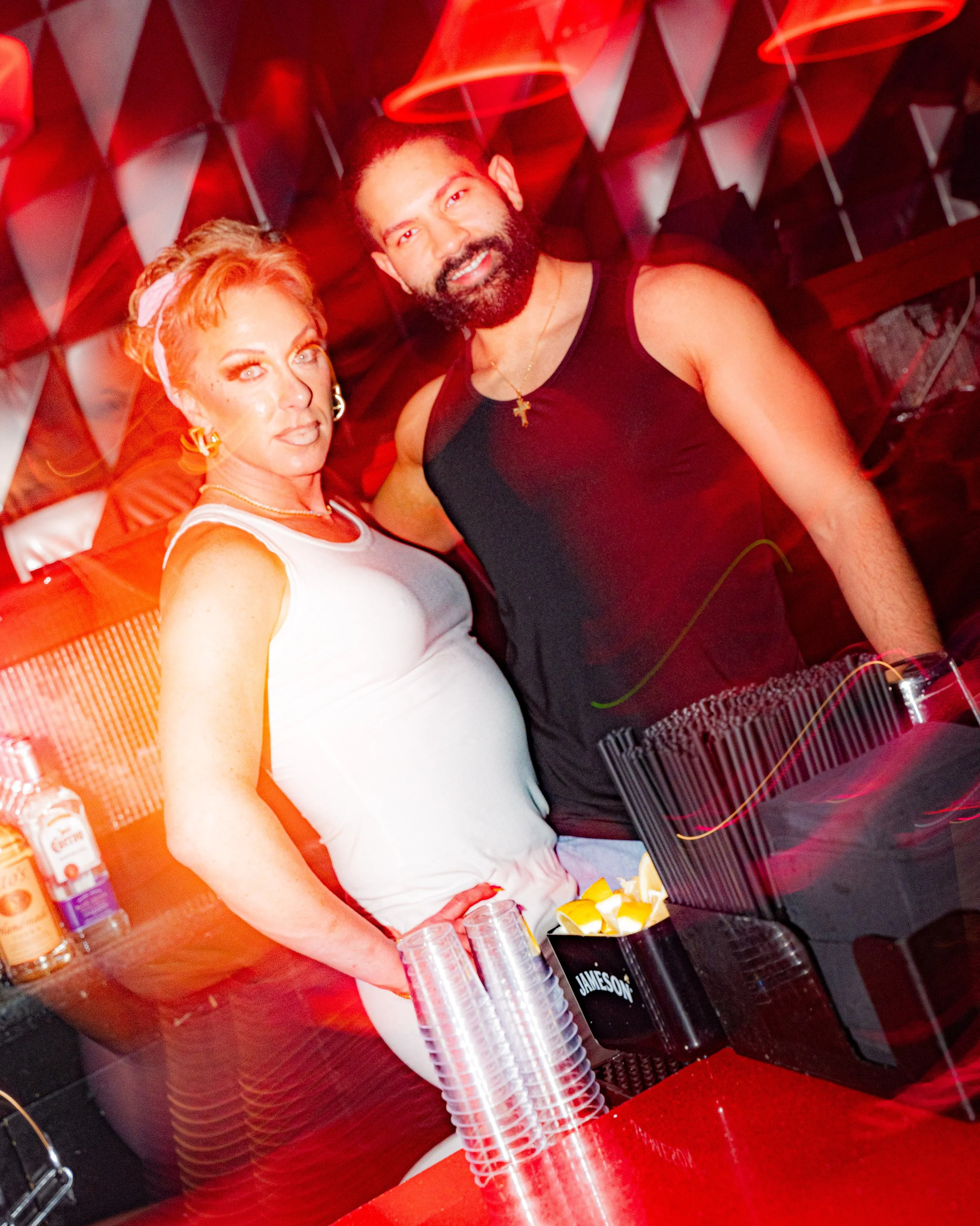 Two people, a woman and a man, standing behind a bar with liquor bottles and plastic cups, with red lighting and party decorations in the background.