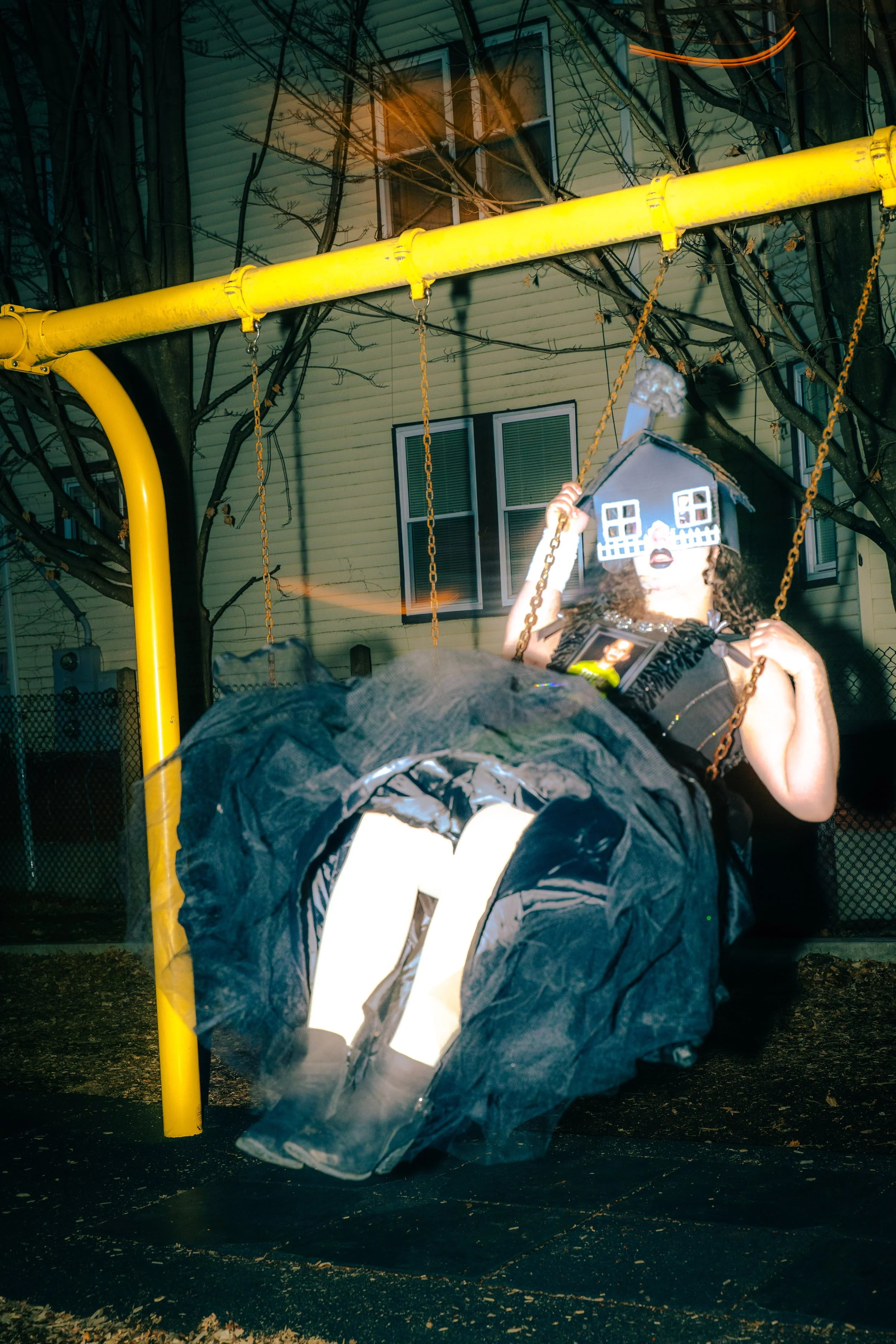 A person dressed in a creative, Halloween costume with a house-shaped mask, sitting on a swing set at night, with a yellow building and leafless trees in the background.