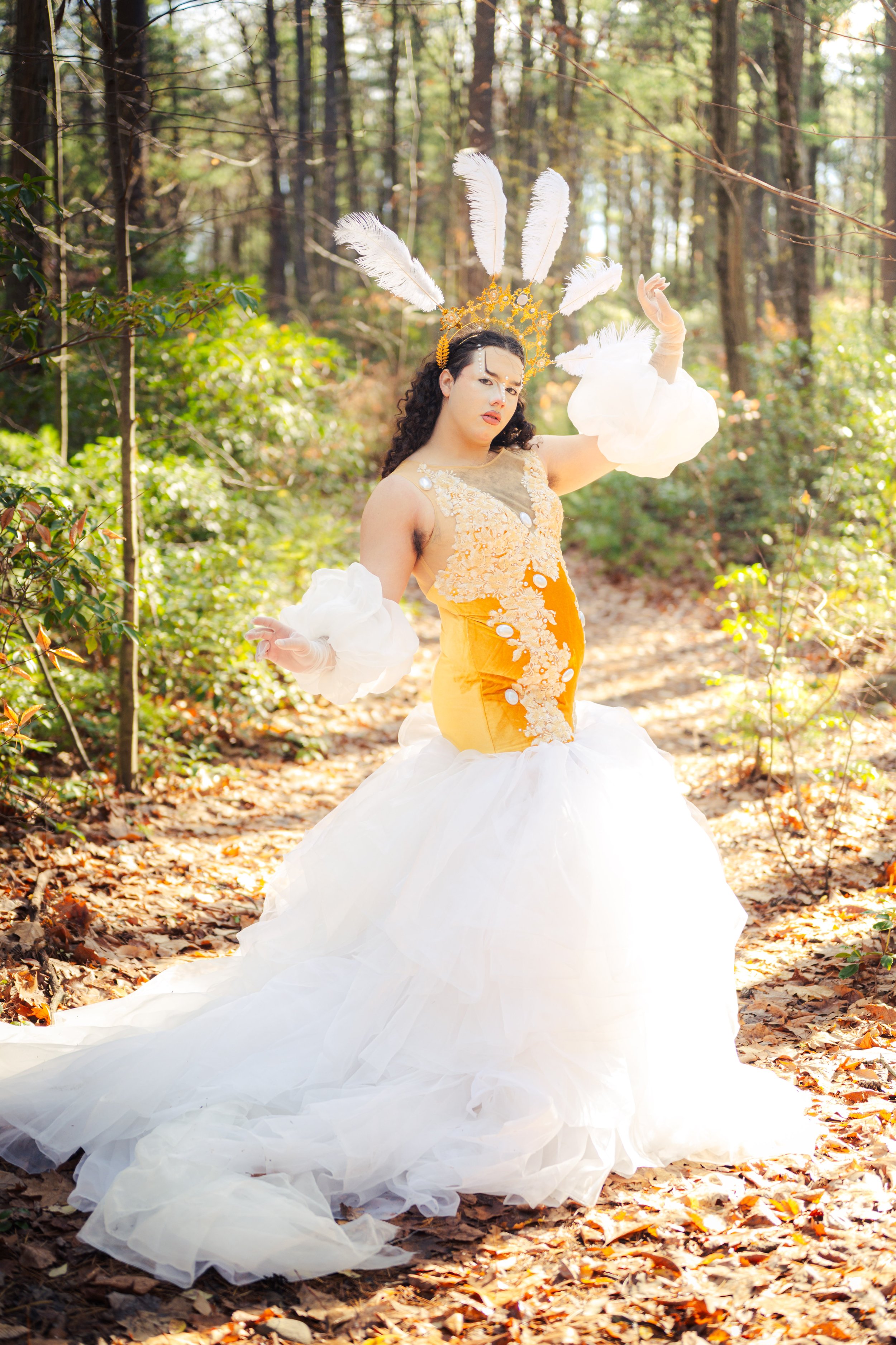 Person dressed in a fantasy costume with a yellow top, white ruffled skirt, and feather crown, posing in a forest.