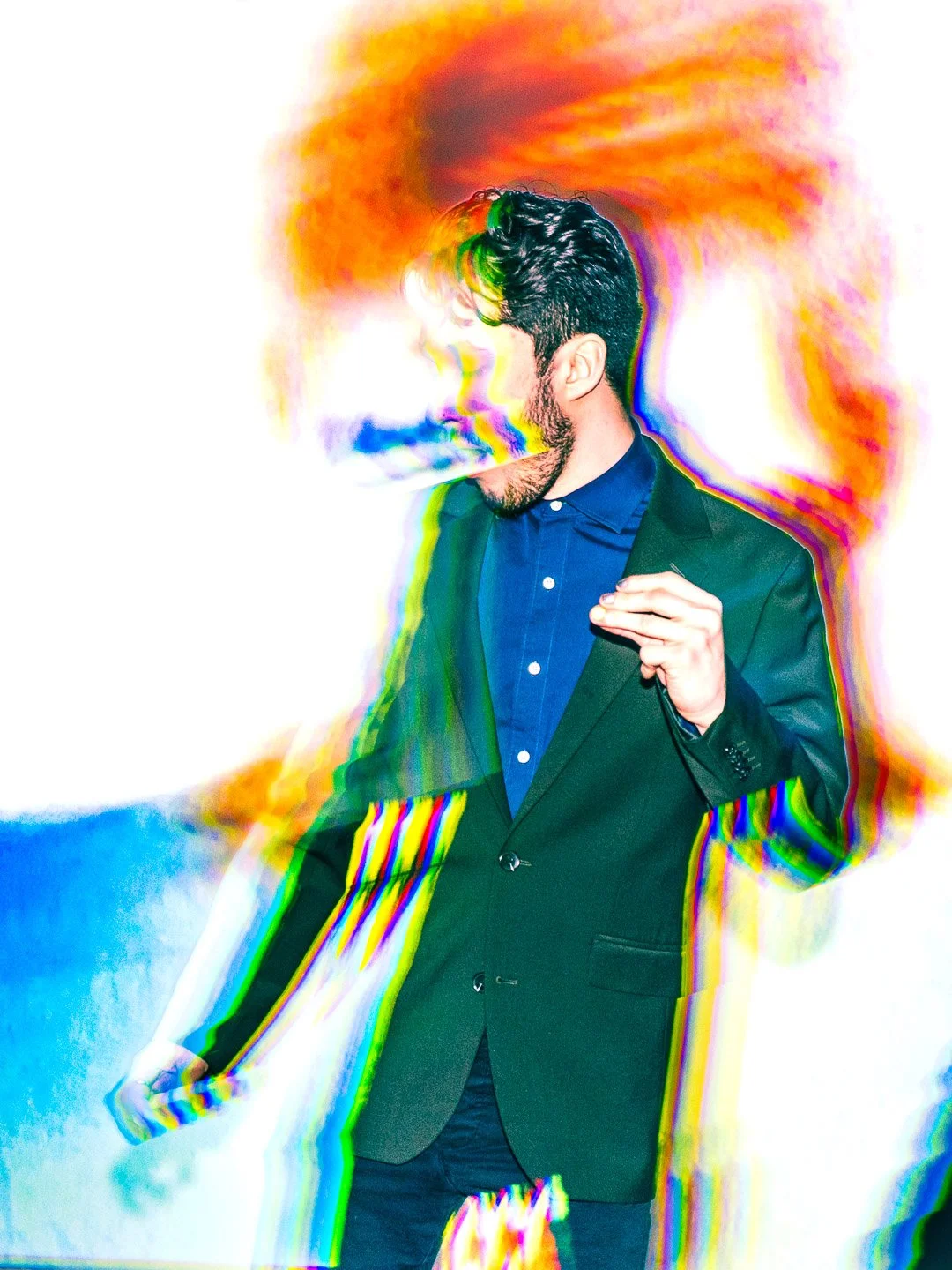 A man in a suit and blue shirt stands against a white background with colorful digital distortion effects, as if his face is dissolving into vibrant rainbow streaks.