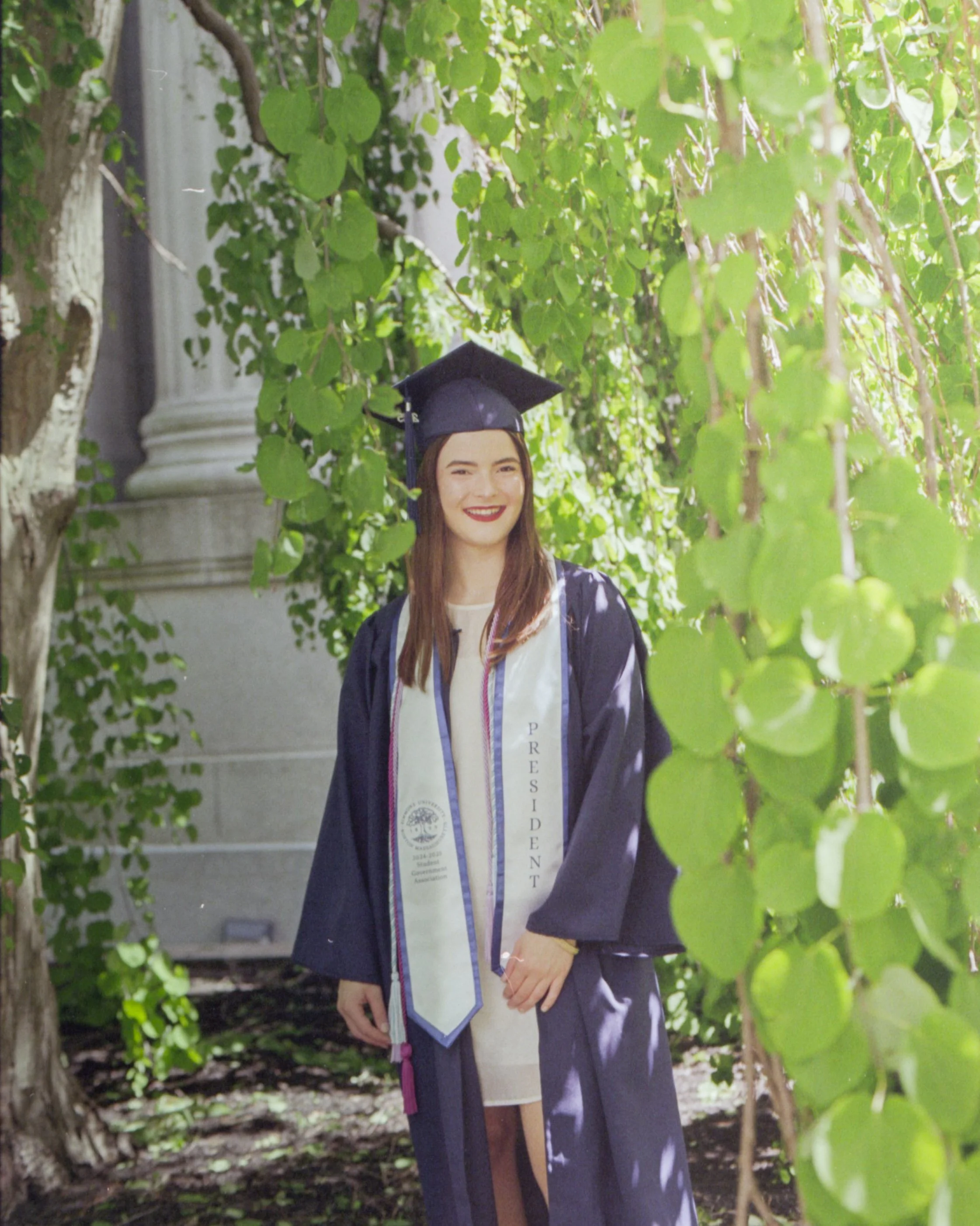 A young woman in a graduation cap and gown with a white stole standing outdoors among green foliage, smiling.