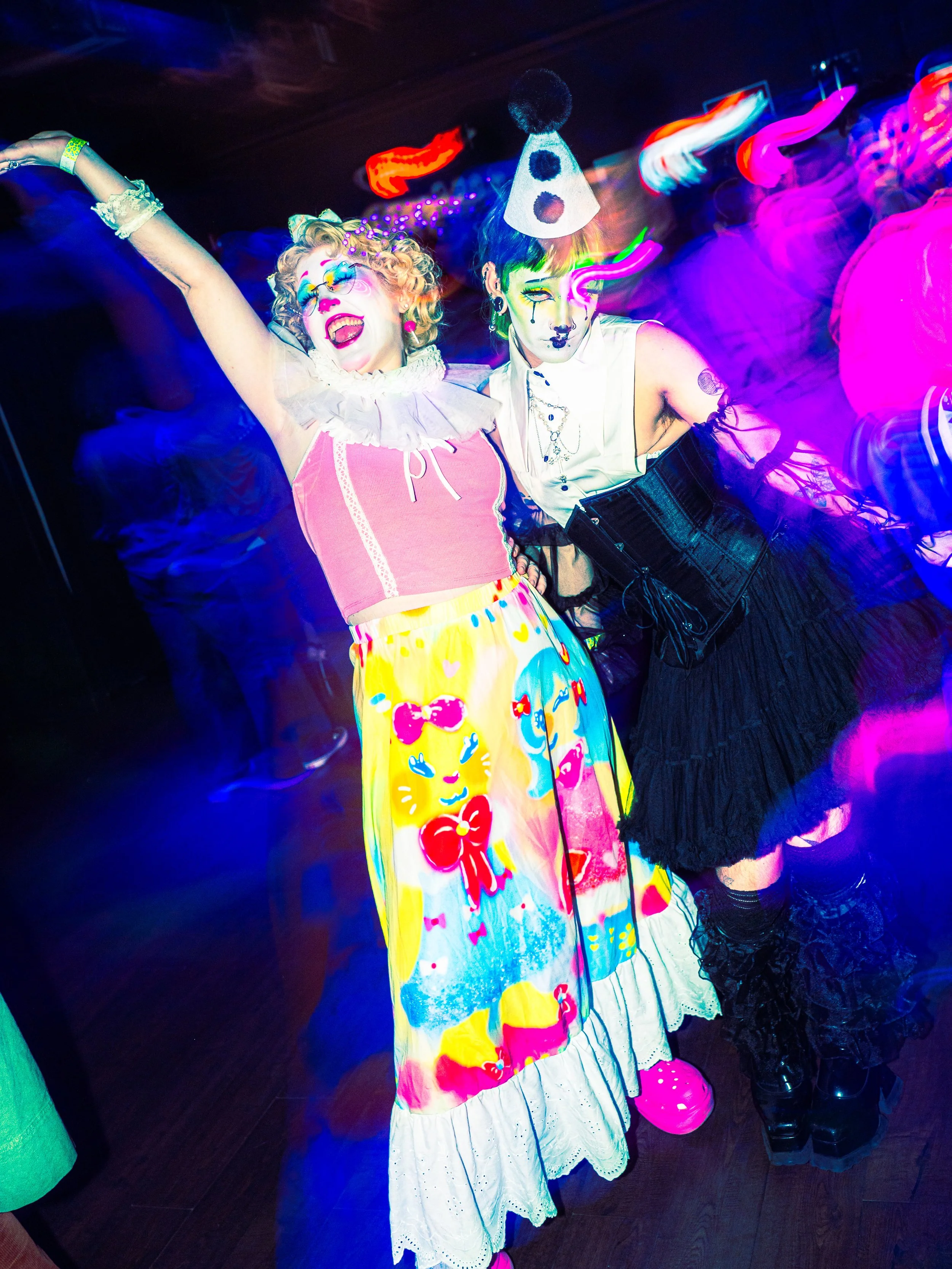 Two people dressed in colorful, eccentric costumes posing at a party with neon lights.
