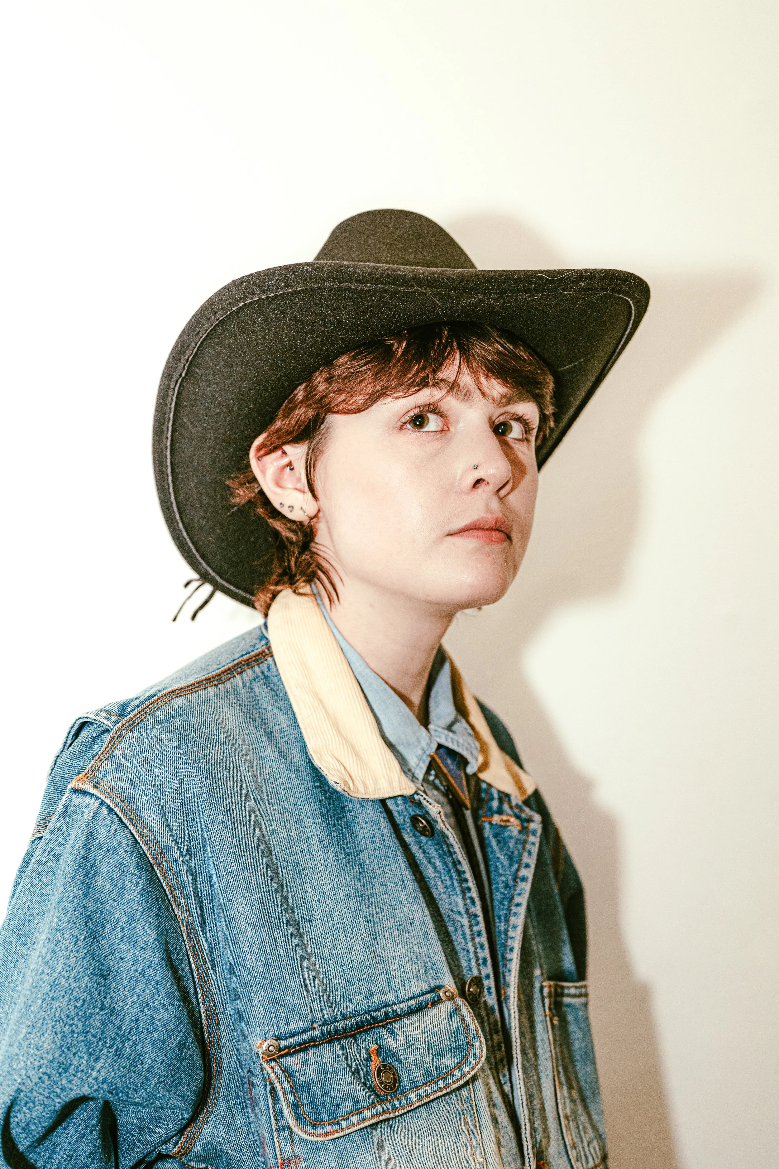 A young person wearing a black cowboy hat and a denim jacket with a beige collar, standing against a white wall.