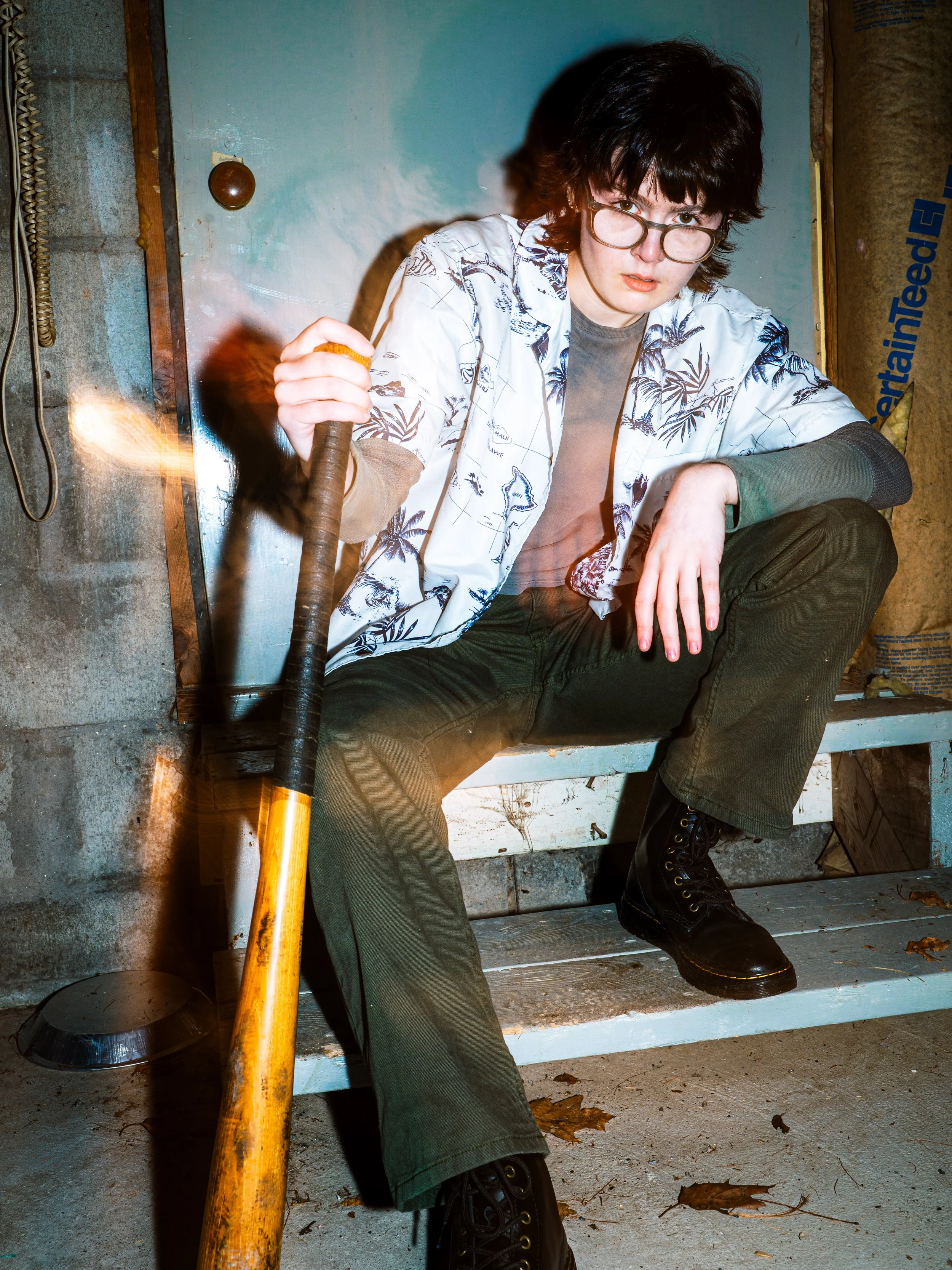 A person with black hair, glasses, a patterned white shirt, dark pants, and black boots sitting on a white wooden bench in a basement-like setting, holding a yellow axe.