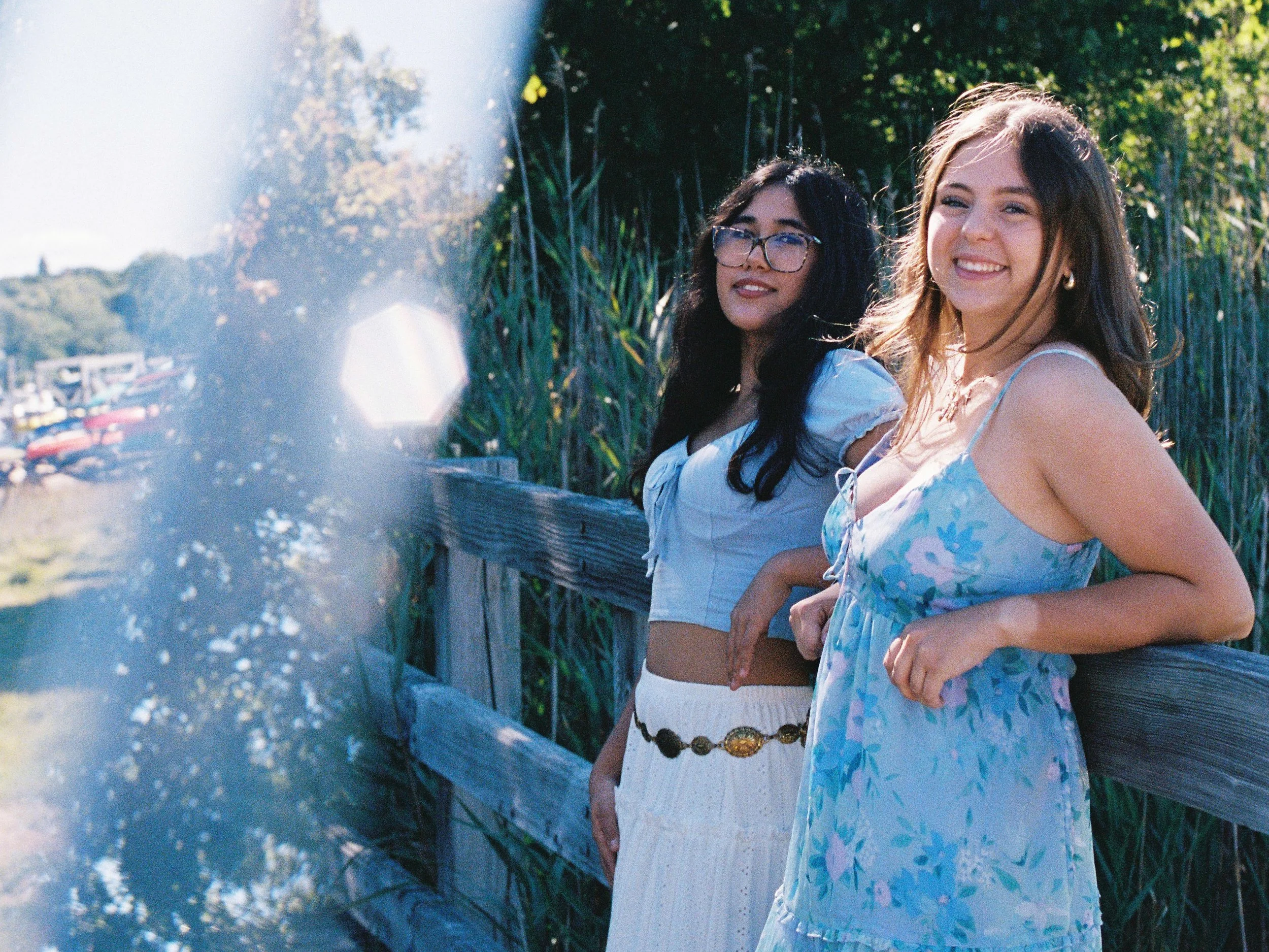 Two young women standing on a wooden pier near tall grass, smiling and looking at the camera, on a sunny day.