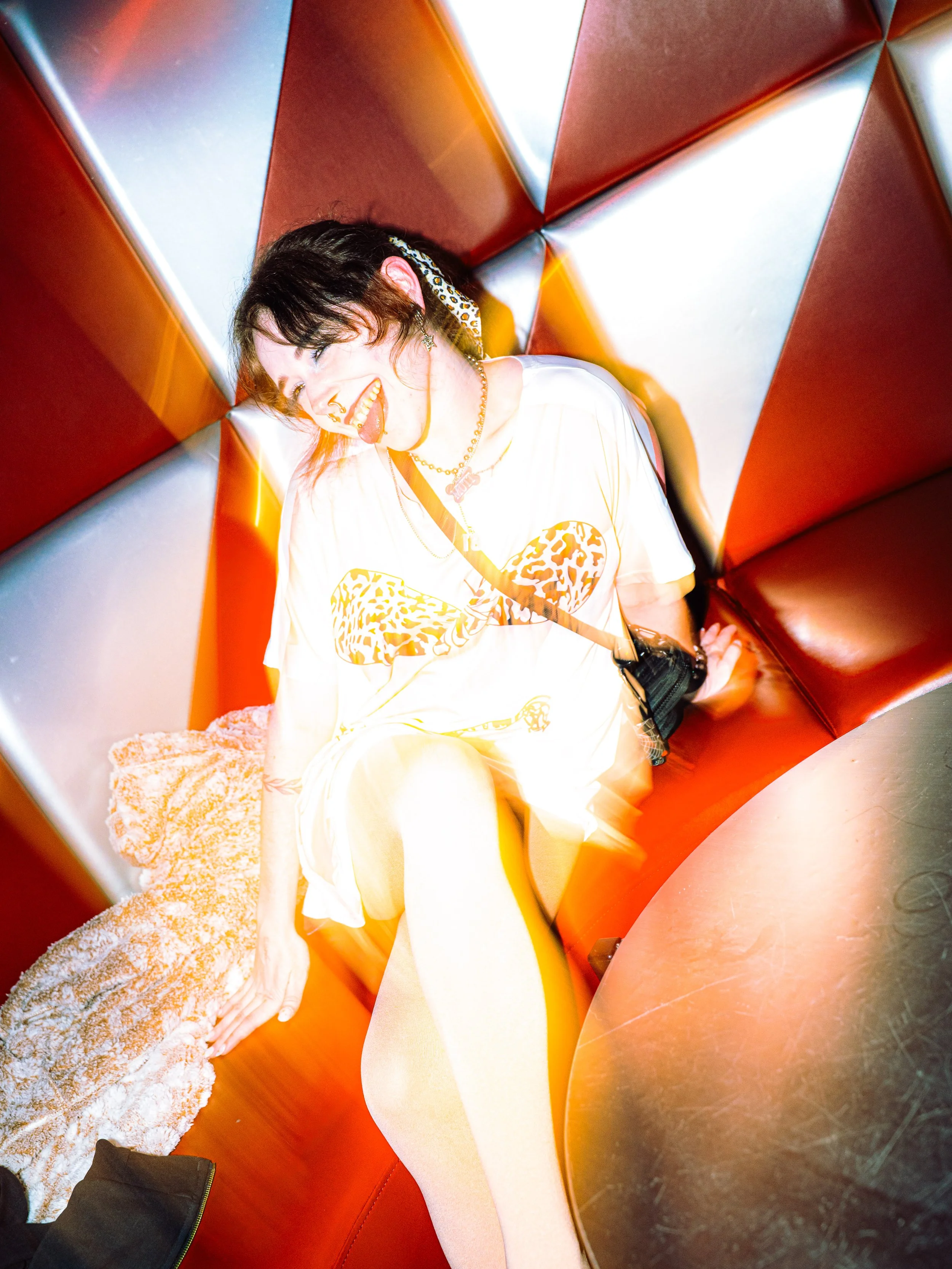 A young woman with short dark hair, wearing a white t-shirt with leopard print heart design, sitting on a red and silver padded booth, smiling and sticking out her tongue.