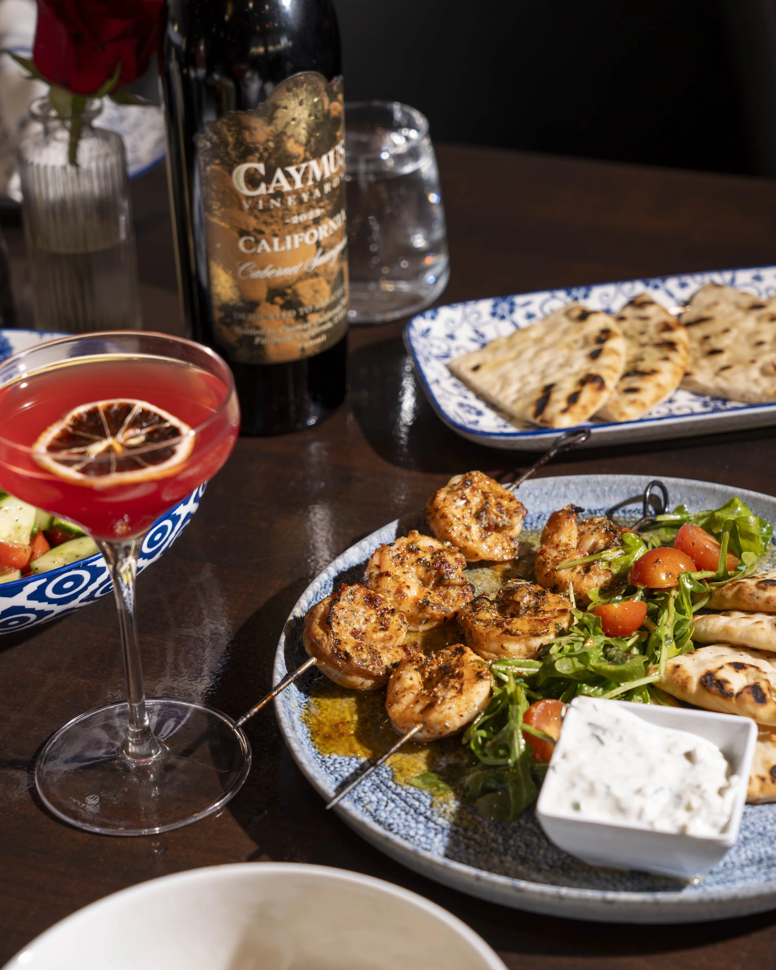 A table with grilled shrimp skewers, fresh salad, flatbread, a glass of red cocktail with lemon slice, a bottle of Caymus wine, and a small dish of white sauce.
