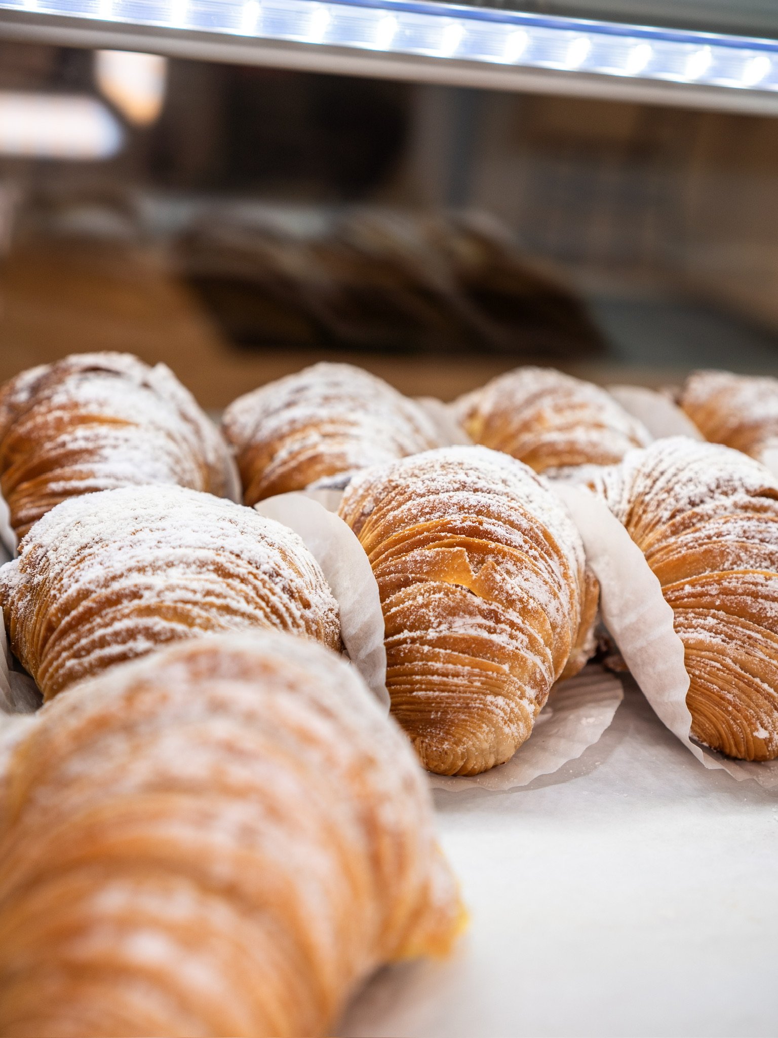 Row of croissants dusted with powdered sugar on baking paper in a bakery display case.