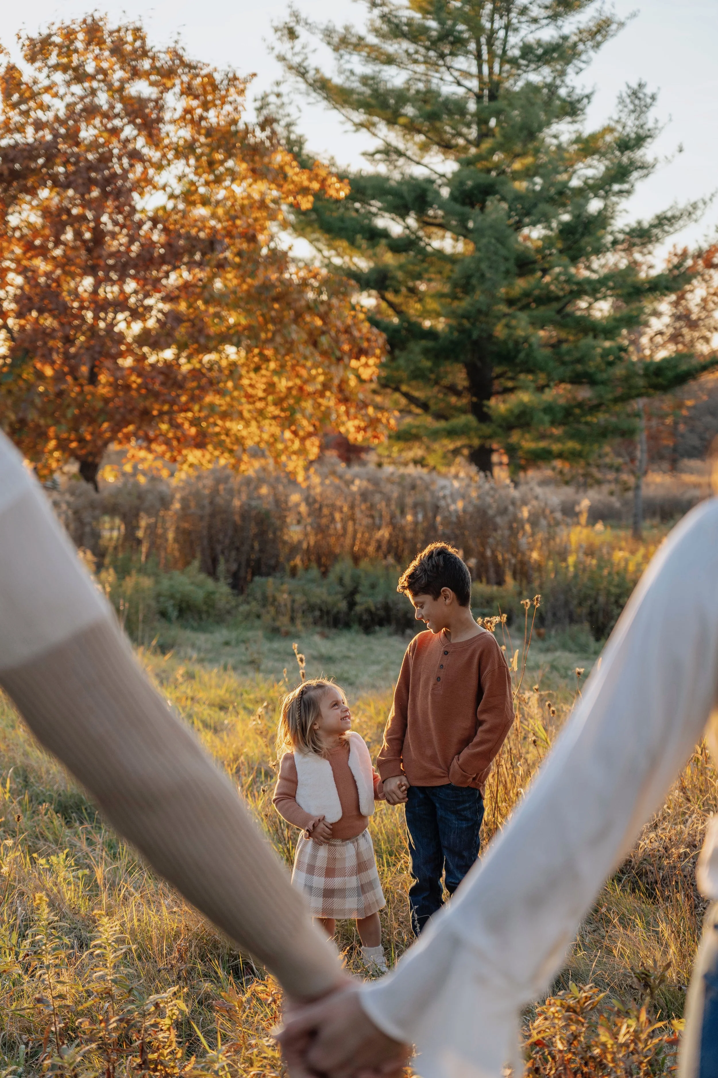 A young girl and a teenage boy hold hands and smile at each other outdoors during sunset, surrounded by autumn trees and grass.