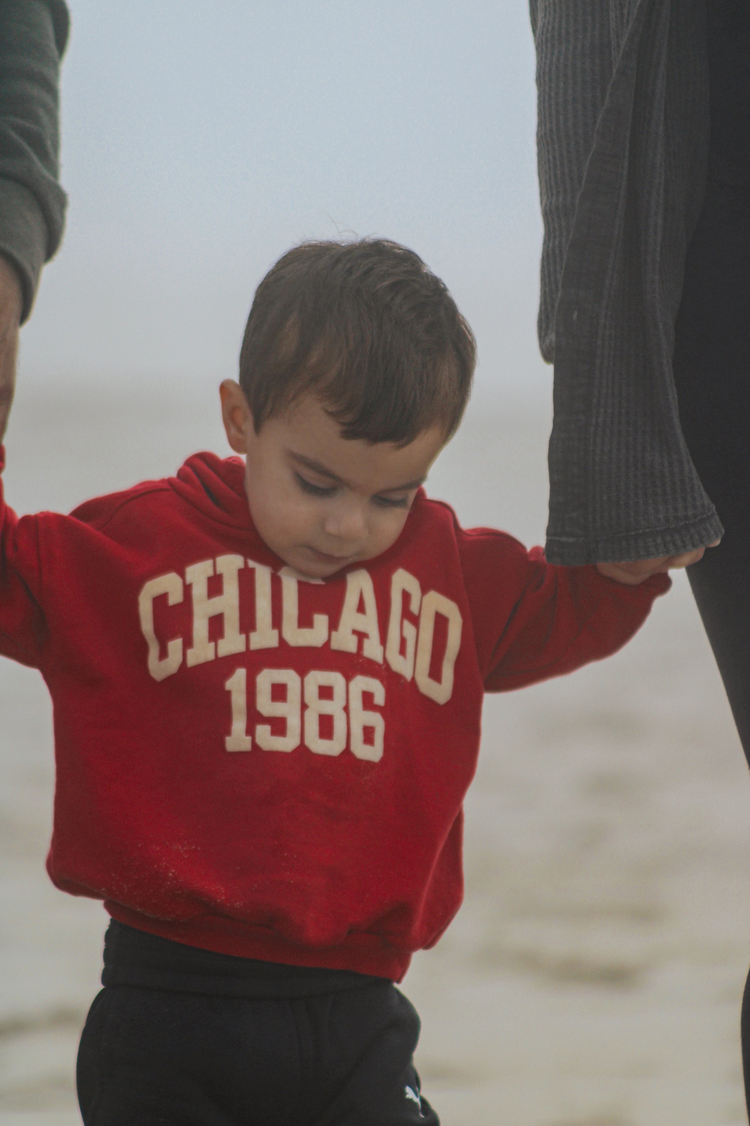 A young boy in a red hoodie with 'CHICAGO 1986' printed on it, looking down and holding hands with two adults, on a beach with a gray sky in the background.