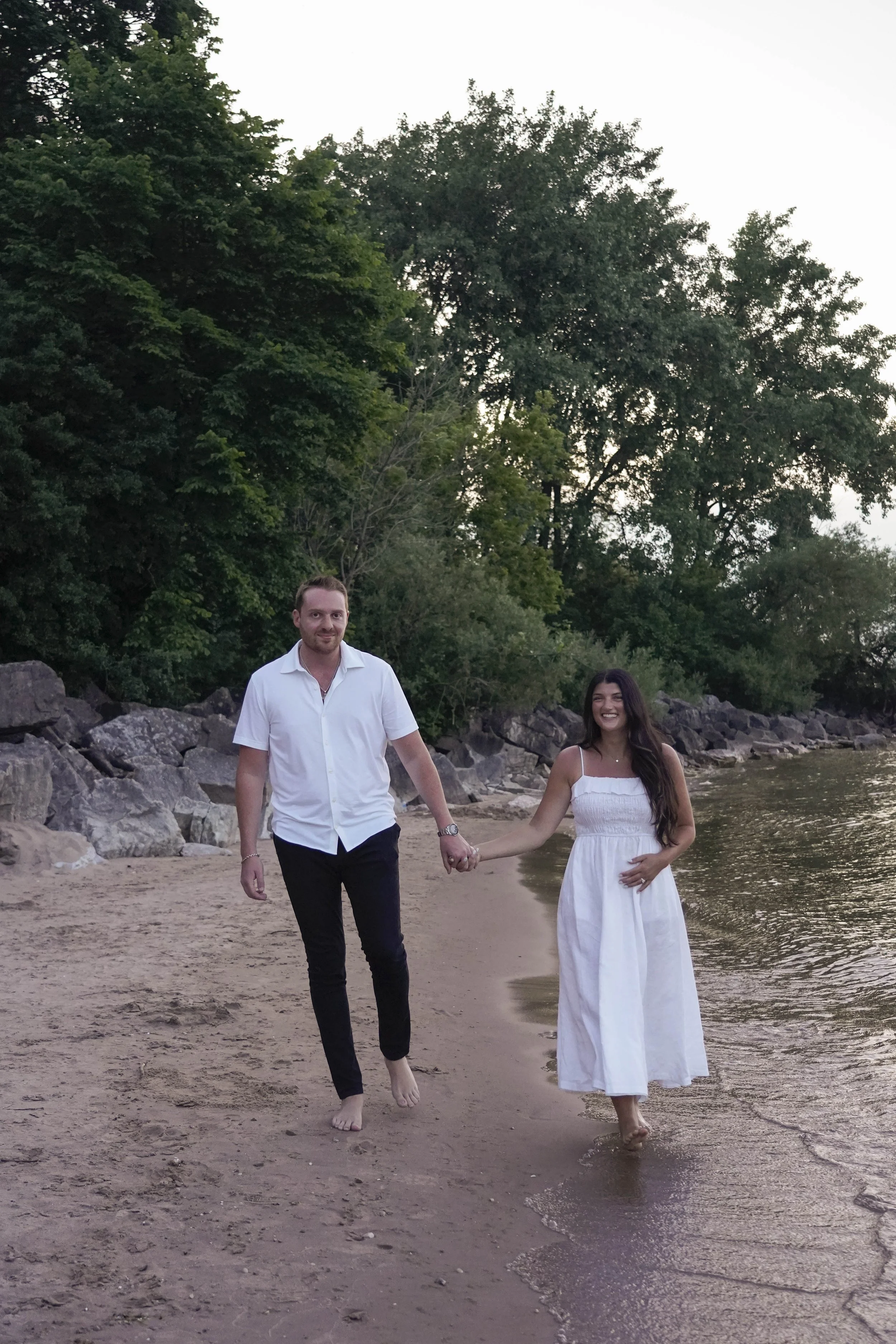A man and woman walking hand in hand along a sandy beach with trees in the background, smiling and enjoying the moment.