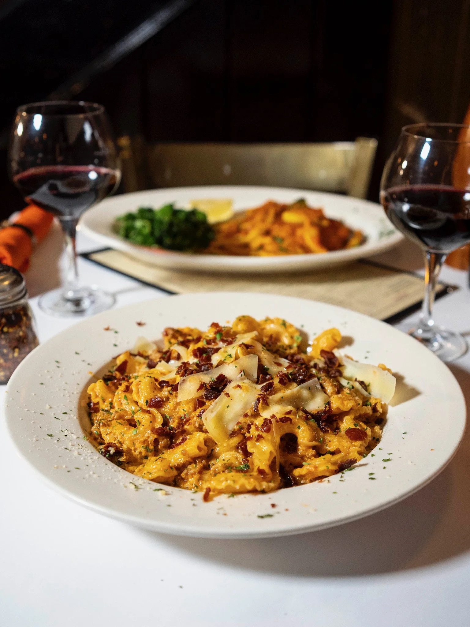 Close-up of a plate of creamy pasta with bacon bits and cheese, with glasses of red wine on a white tablecloth, and another plate of pasta in the background.