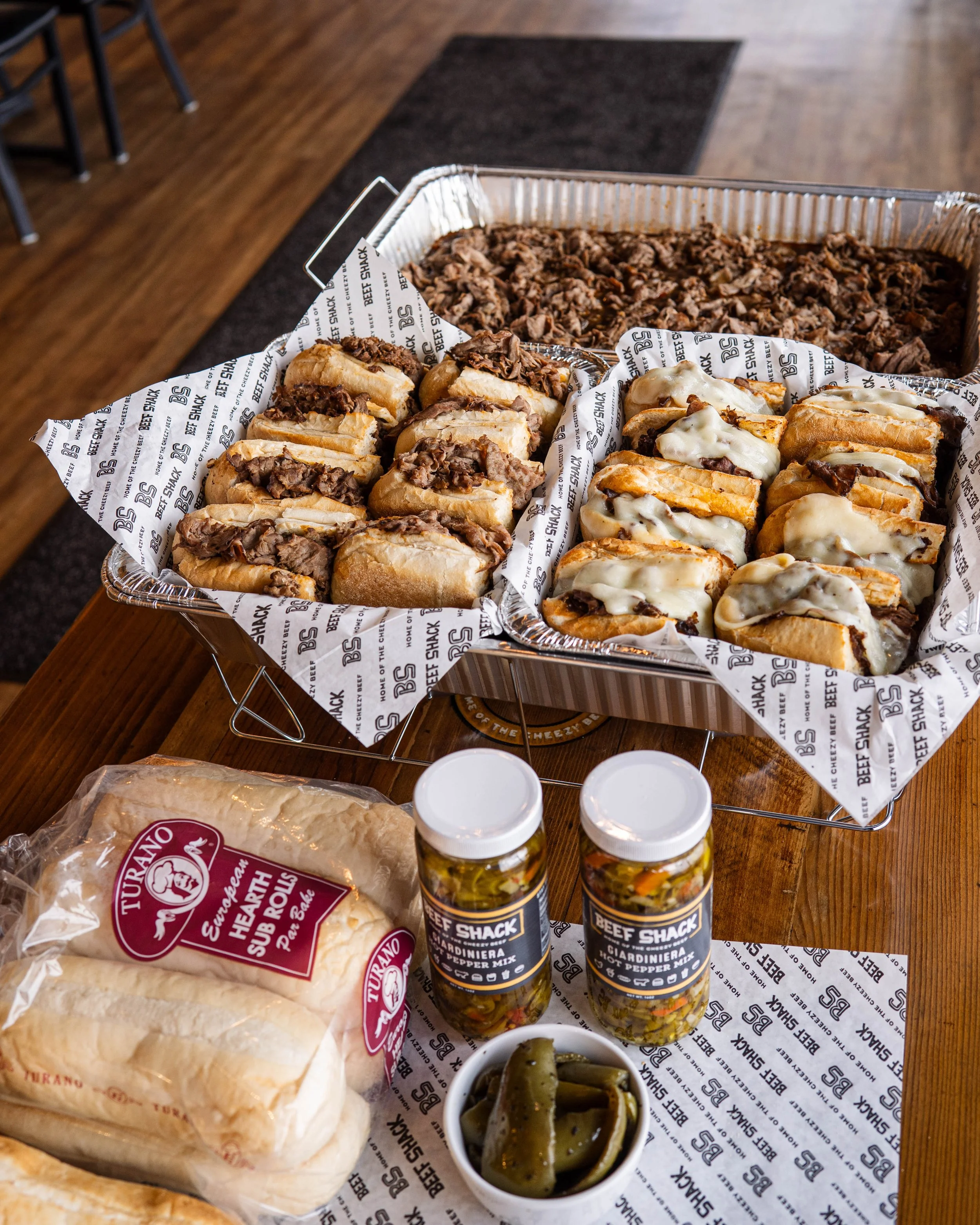 A tray of beef sandwiches with shredded beef, some topped with melted cheese, and a large tray of shredded beef, with bottles of pickled peppers, a container of pickled jalapeños, and a bag of bread rolls on a wooden table.