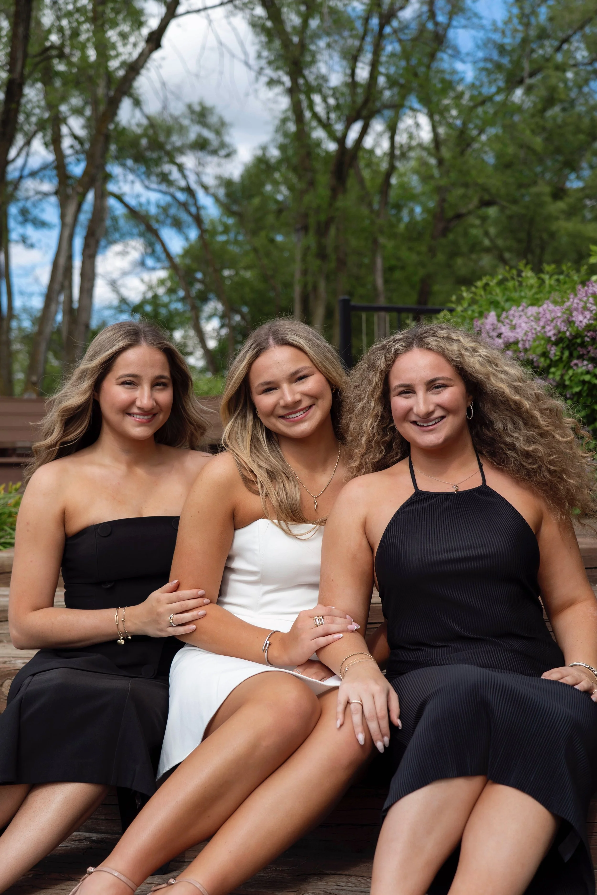 Three women sitting on a wooden bench outdoors on a sunny day, surrounded by trees and flowering bushes.