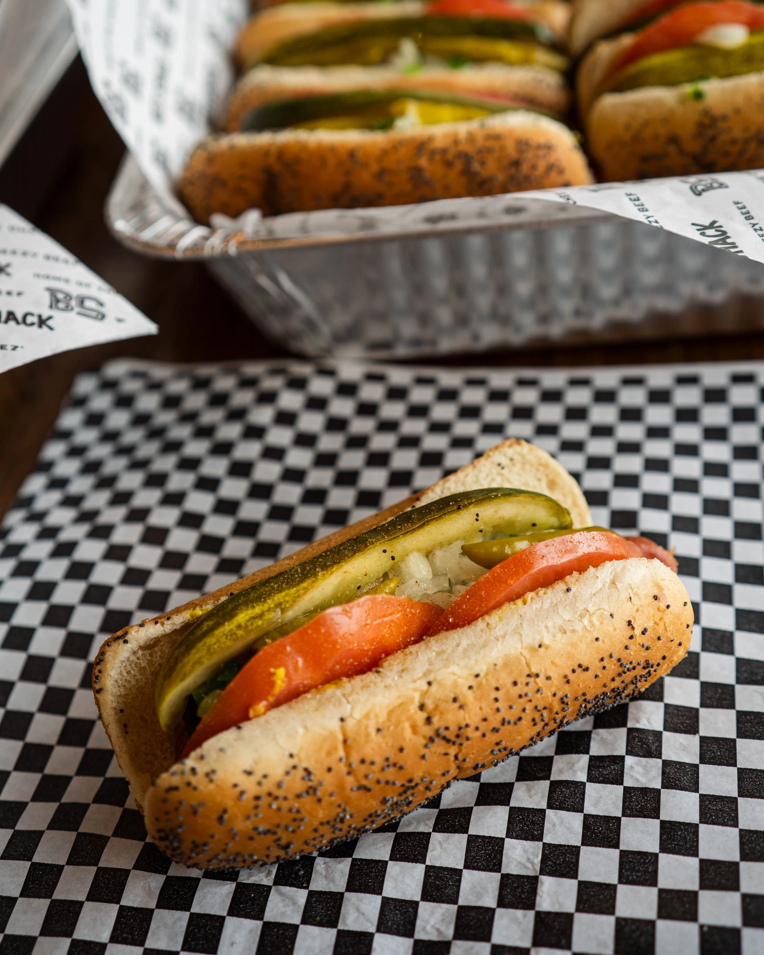 A hot dog in a poppy seed bun with pickles, tomato slices, onion, and jalapeño peppers, placed on checkered paper with a tray of more hot dogs in the background.