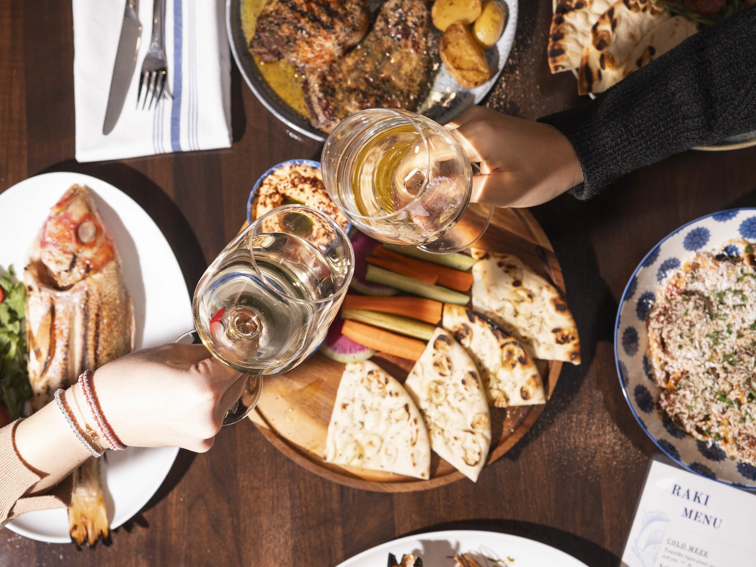People raising glasses of white wine over a table with various dishes, including grilled meat, vegetables, pizza, rice, and bread.