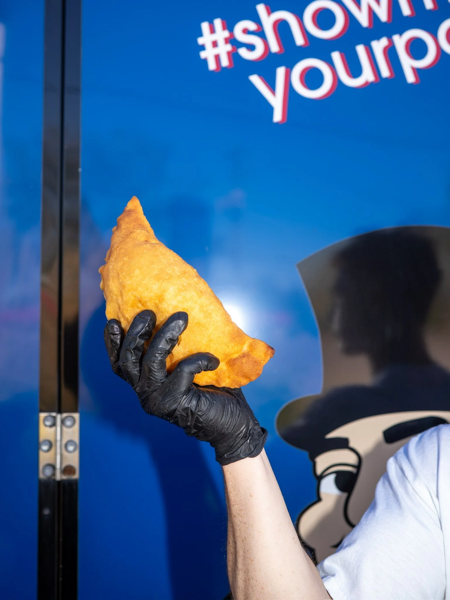 A person wearing a black glove is holding a fried empanada in front of a blue wall with the hashtag '#showingyour' partially visible.