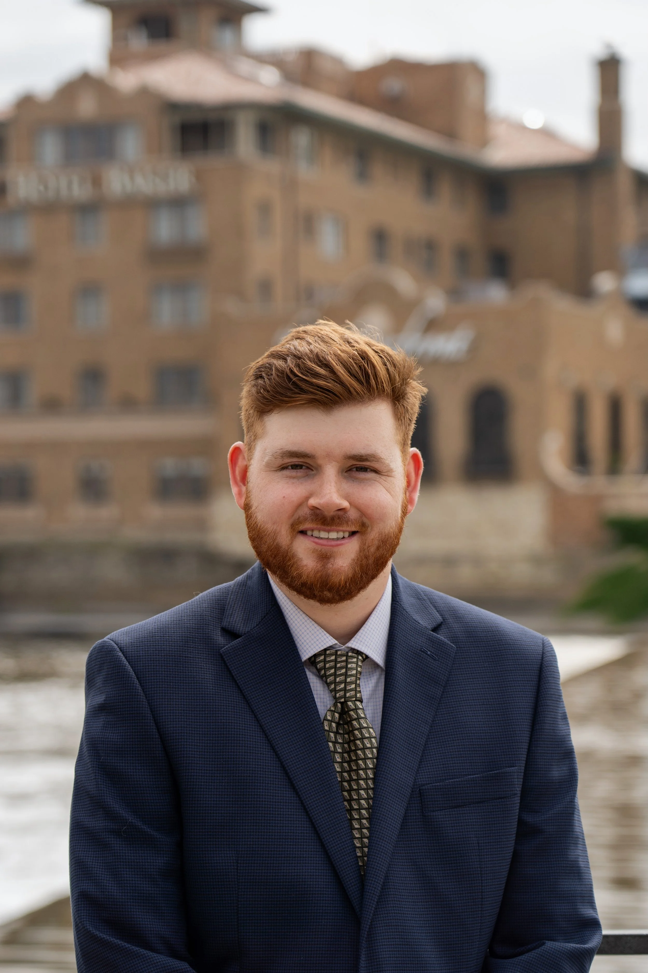 A man in a blue suit and tie standing outdoors near water with a historic brick building in the background.
