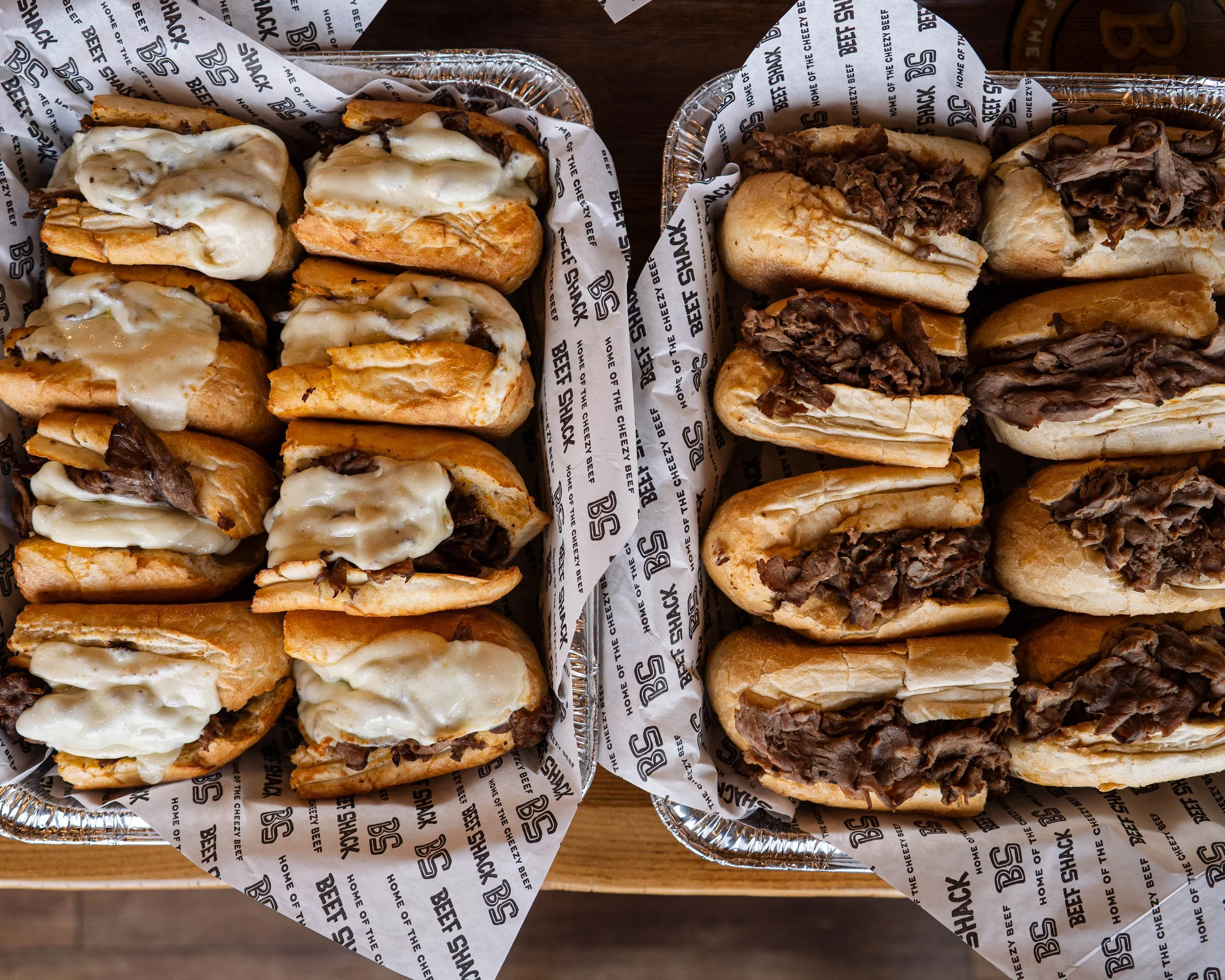Two aluminum trays filled with small sandwiches, one with melted cheese and beef, and the other with shredded beef, on a wooden surface.