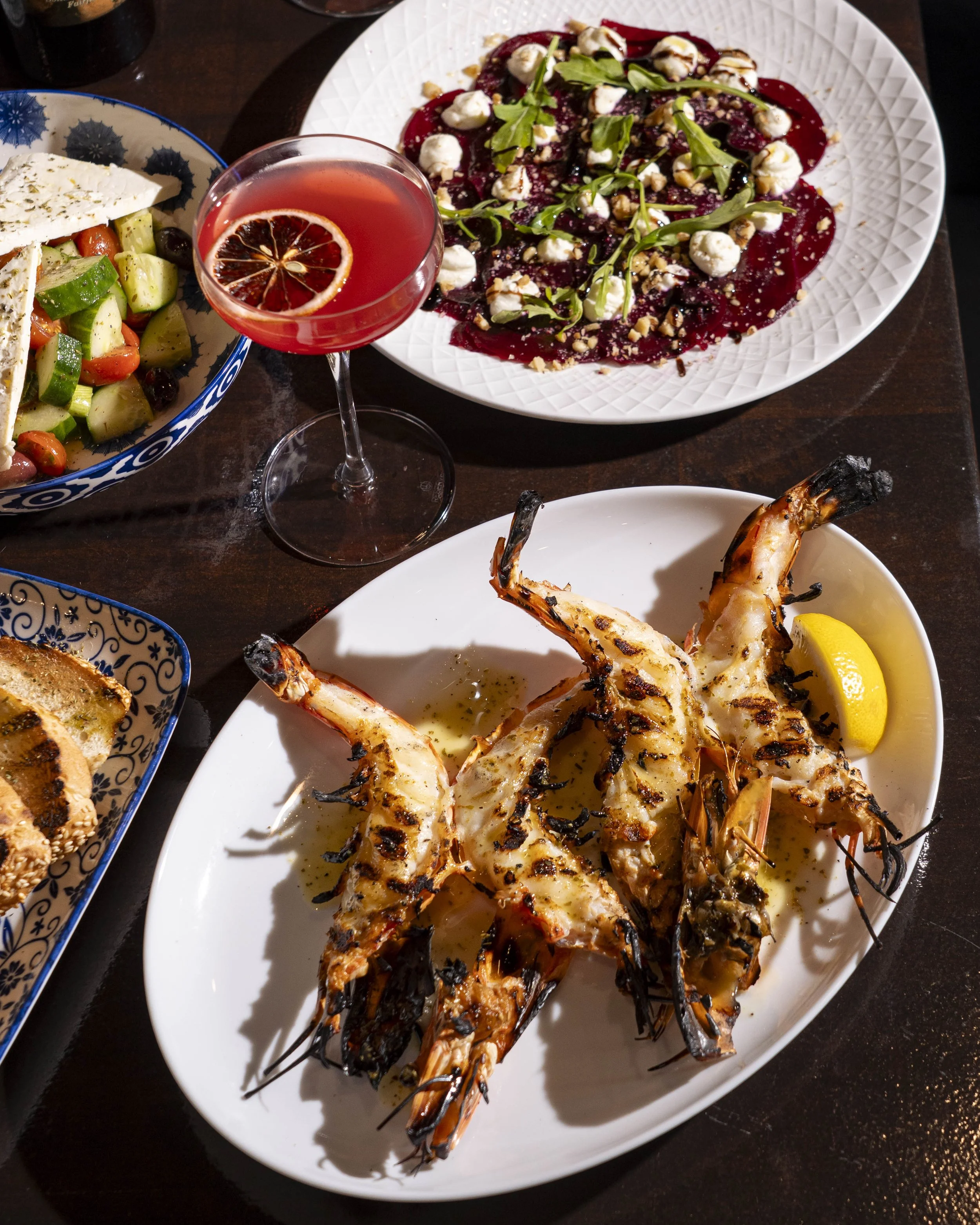 Grilled shrimp with lemon wedge on a white platter, salad with cucumbers, tomatoes, and cheese on a blue and white patterned dish, a beet salad with greens, ricotta cheese, and nuts on a white plate, and a cocktail with a lemon slice