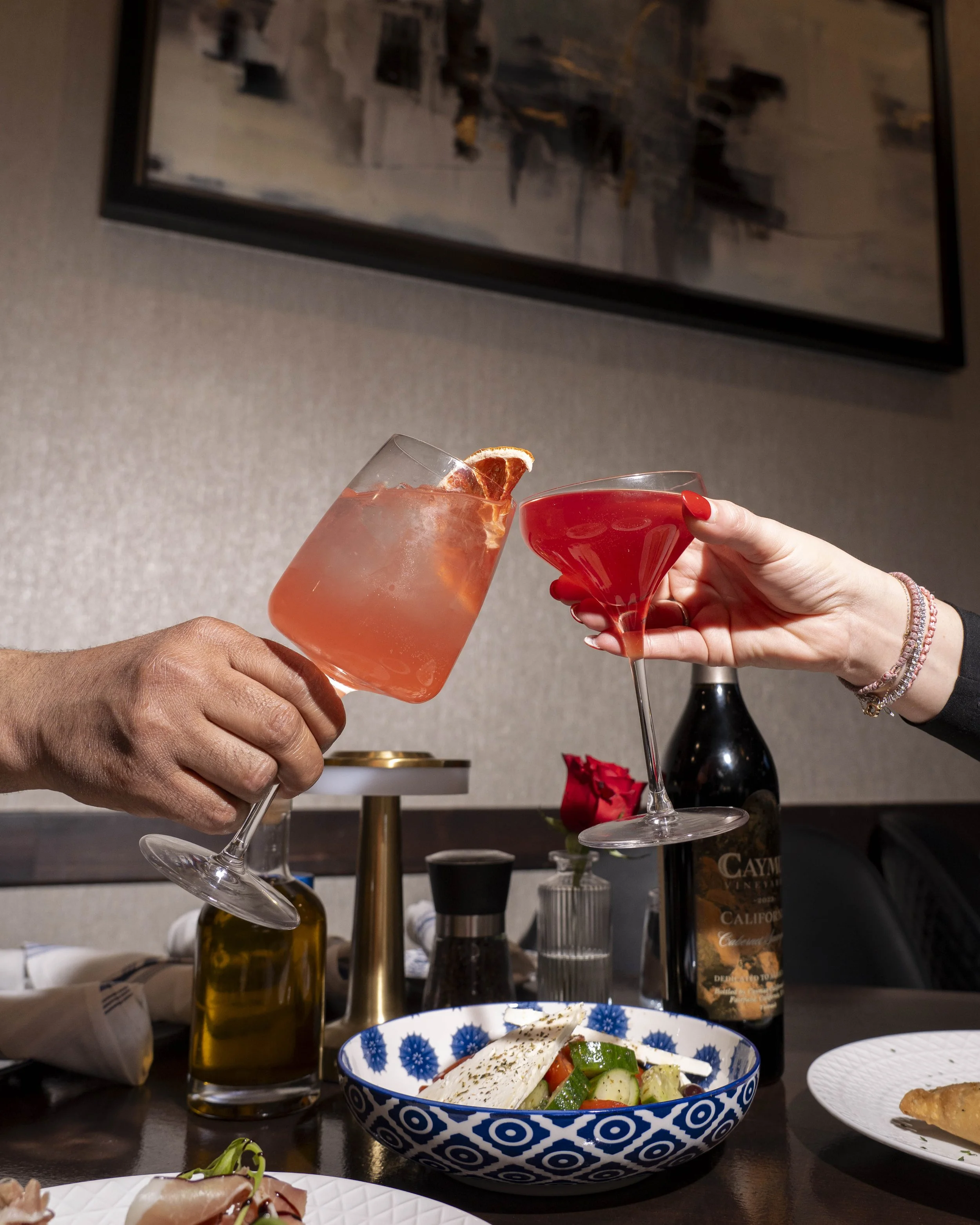 Two people clinking glasses, one with a pink cocktail topped with a slice of dried citrus, and the other with a red cocktail in a martini glass, on a table with a salad, wine bottle, and other dishes.