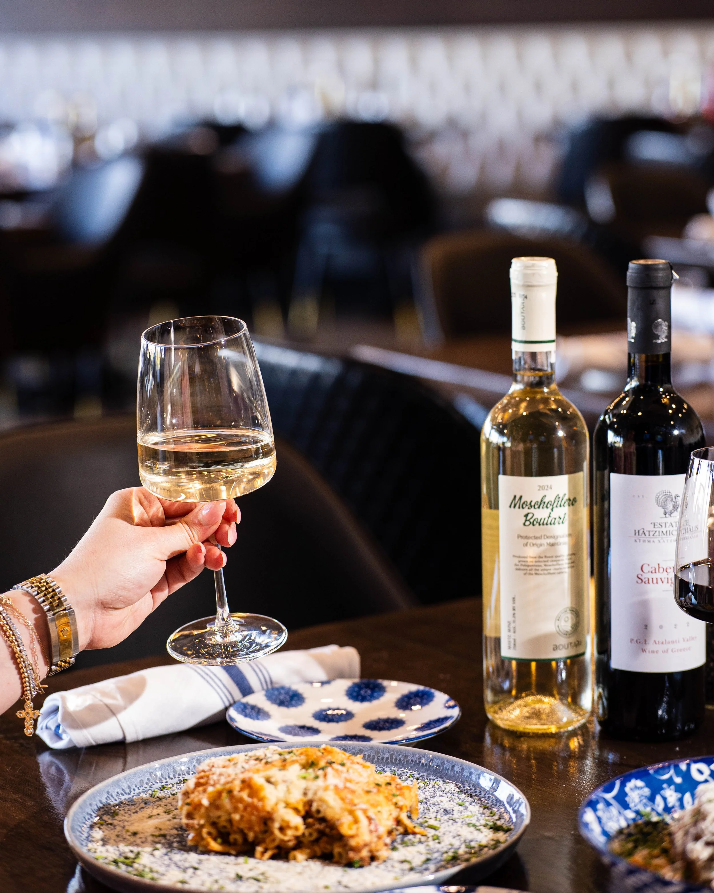 Person holding a glass of white wine at a dining table with plates of food, wine bottles, and a napkin.
