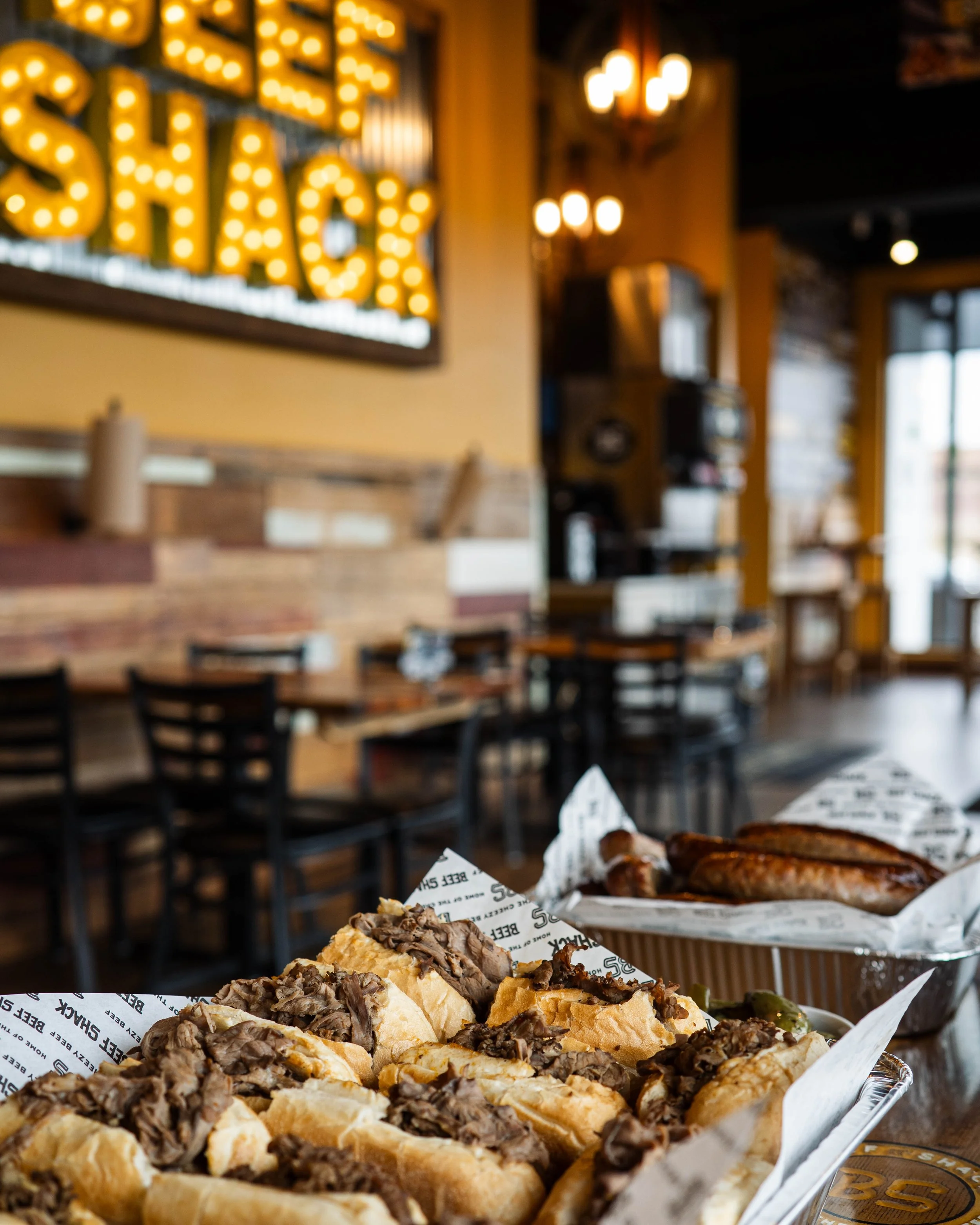 French fries topped with shredded beef served with grilled sausage in a restaurant with yellow walls, wooden decor, and a brightly lit sign that says 'EAT MY SHACK' in the background.