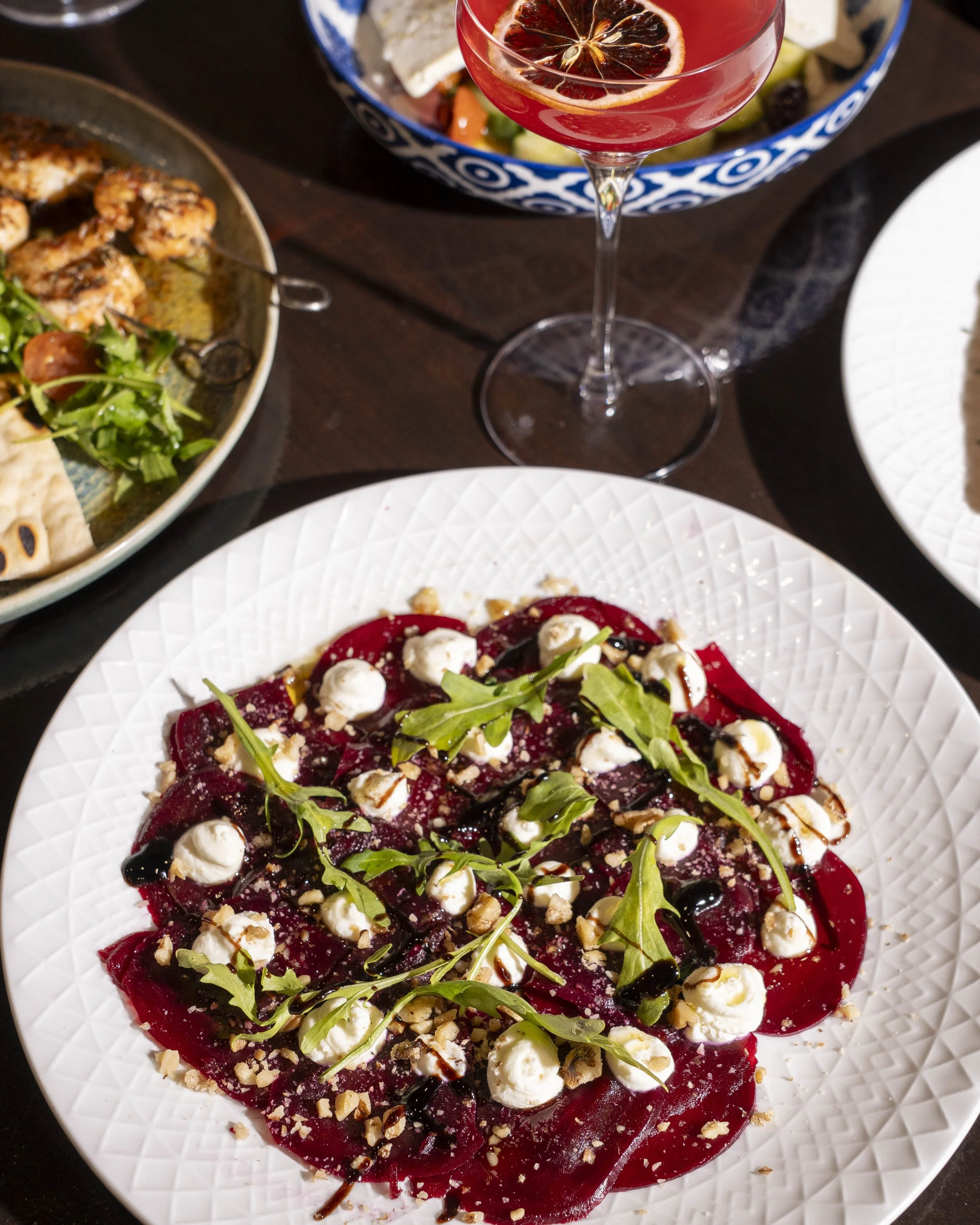 Plate of beet carpaccio topped with goat cheese, arugula, chopped nuts, balsamic glaze, and black pepper, with a glass of red cocktail garnished with a blood orange slice in the background.