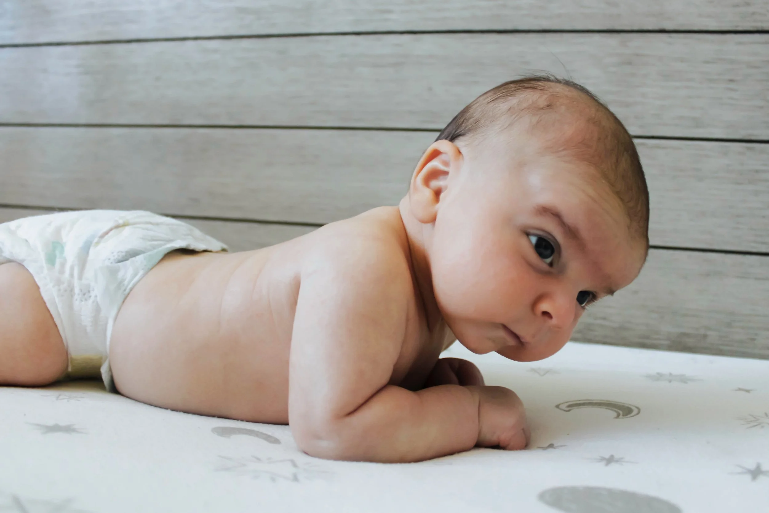 A young baby lying on stomach on a bed with a light-colored sheet featuring star and moon designs, with a wooden-paneled wall in the background.