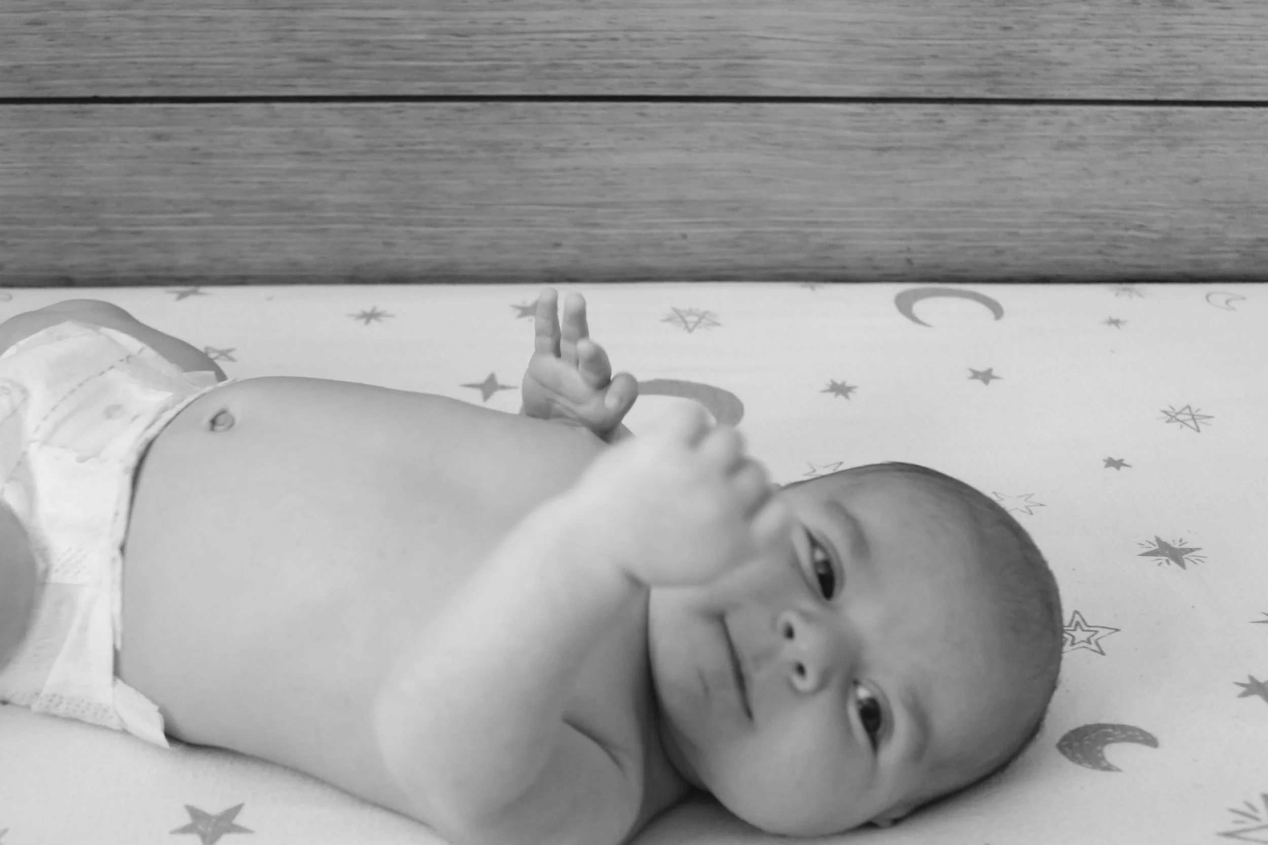 A baby lying on a patterned sheet, with a wooden wall in the background, making a peace sign with one hand and looking at the camera.