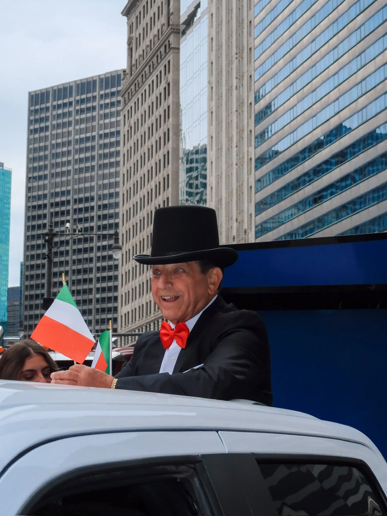 A man dressed in a tuxedo with a red bow tie and a tall black hat riding in a vehicle during a parade, holding an Irish flag. Tall modern buildings in the background suggest an urban setting.
