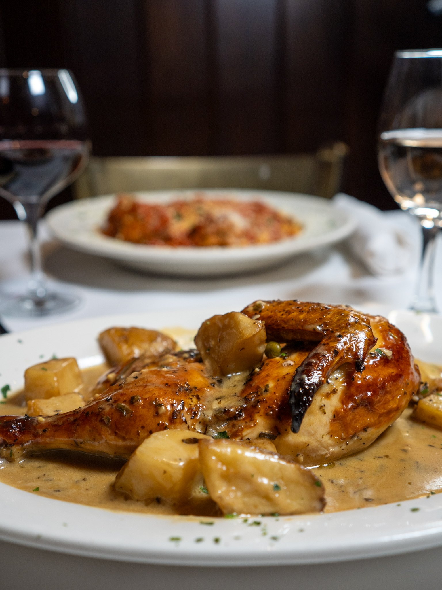 A plate with roasted chicken topped with sauce, surrounded by potatoes, served on a white tablecloth in a restaurant setting.